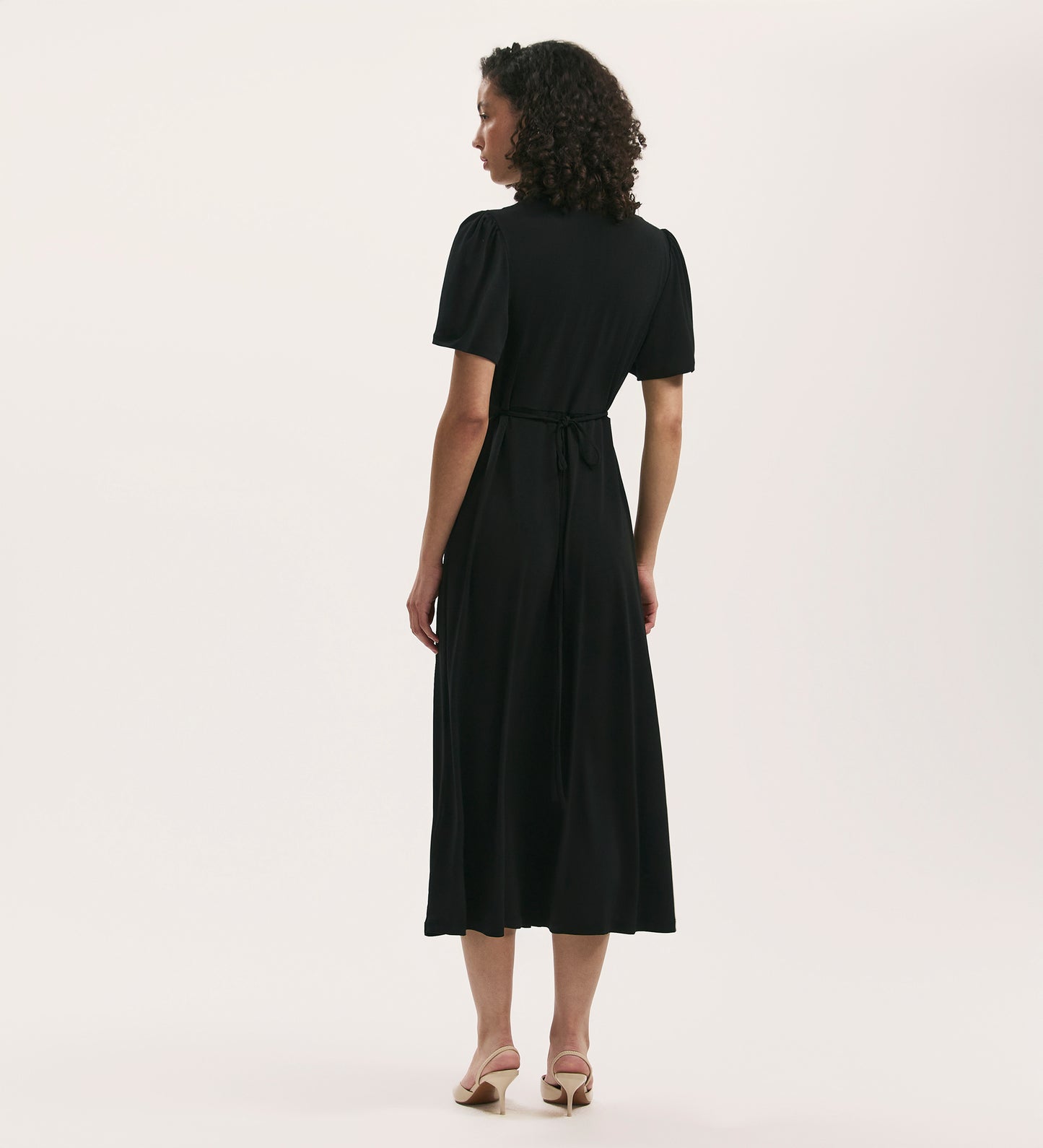 A person with curly hair, seen from behind, wears the April Black Jersey Midi Dress—a relaxed-fit, tie-waist midi in jersey fabric—paired with beige heeled sandals against a plain light background.