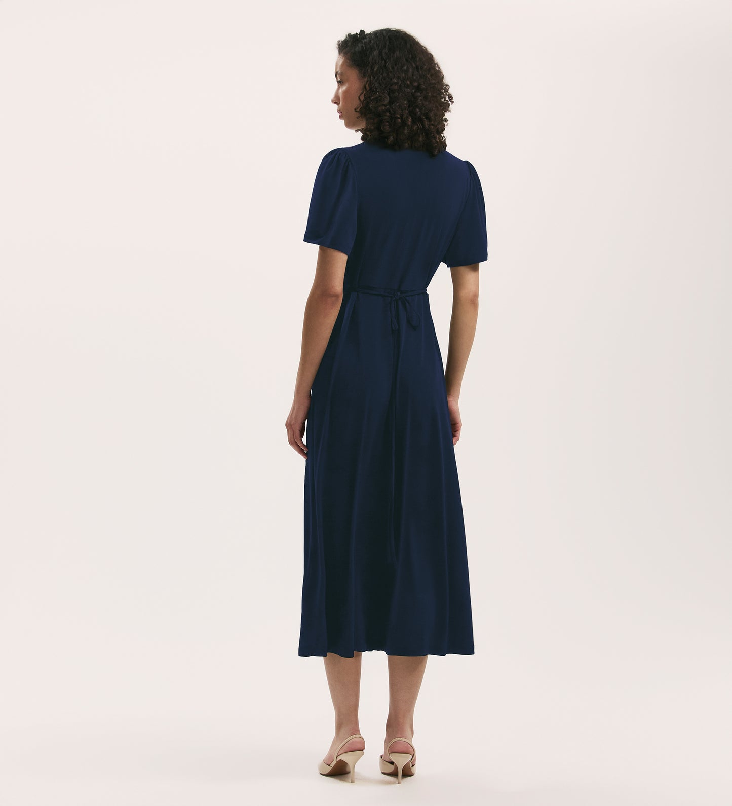 A woman with curly hair, standing and facing away, wears the April Navy Jersey Midi Dress—relaxed fit with short sleeves and a waist tie—paired with light heels against a plain light background.