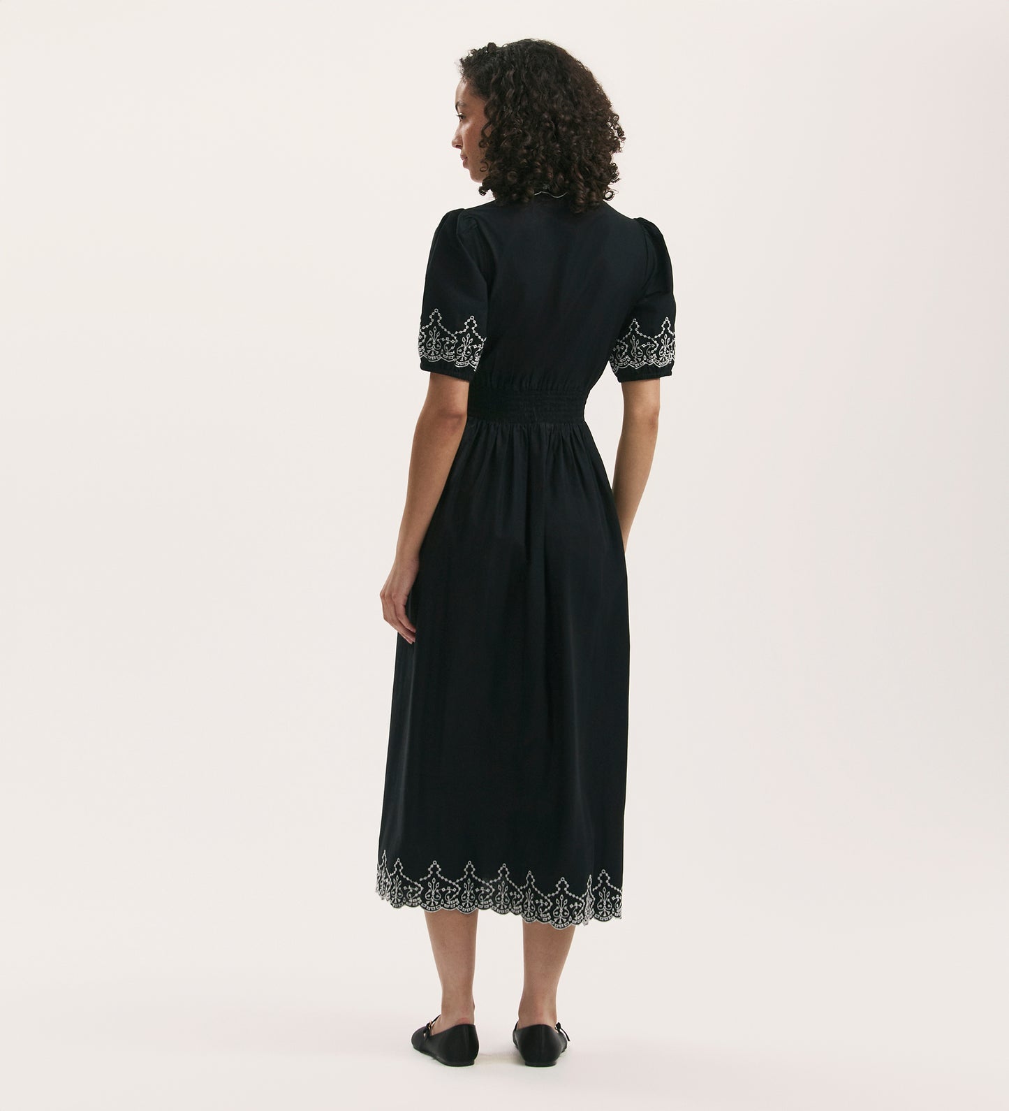 A woman with curly hair faces away, wearing the Arianna Black Embroidered Cotton Midi Dress featuring short puff sleeves and white embroidery on the sleeves and hem, paired with black flat shoes.