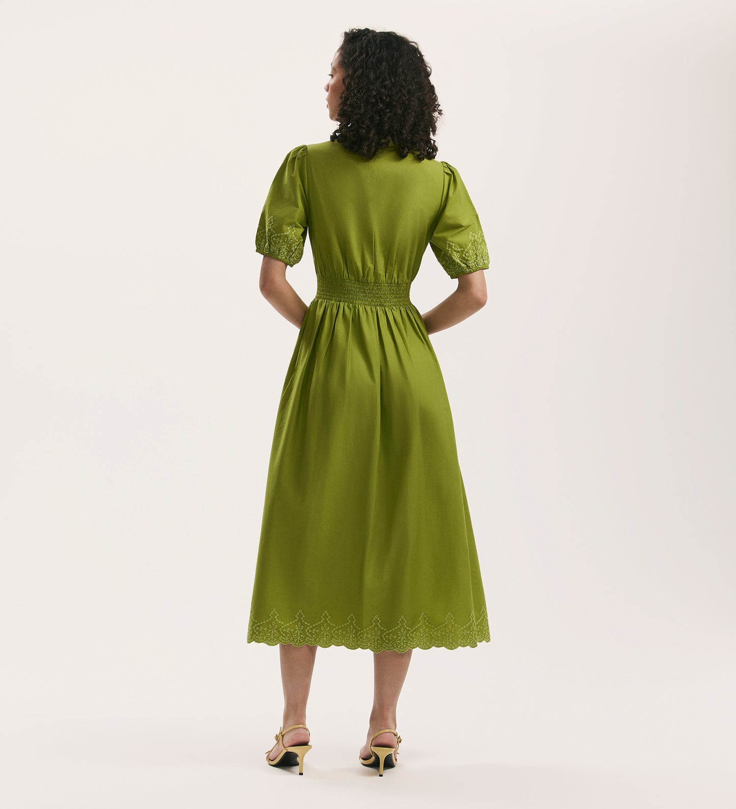 A woman faces away, wearing the Arianna Light Green Embroidered Cotton Midi Dress with short sleeves, fitted waist, lace-trimmed sleeves and hem, scalloped edges, embroidery details, and paired with open-toe heels.