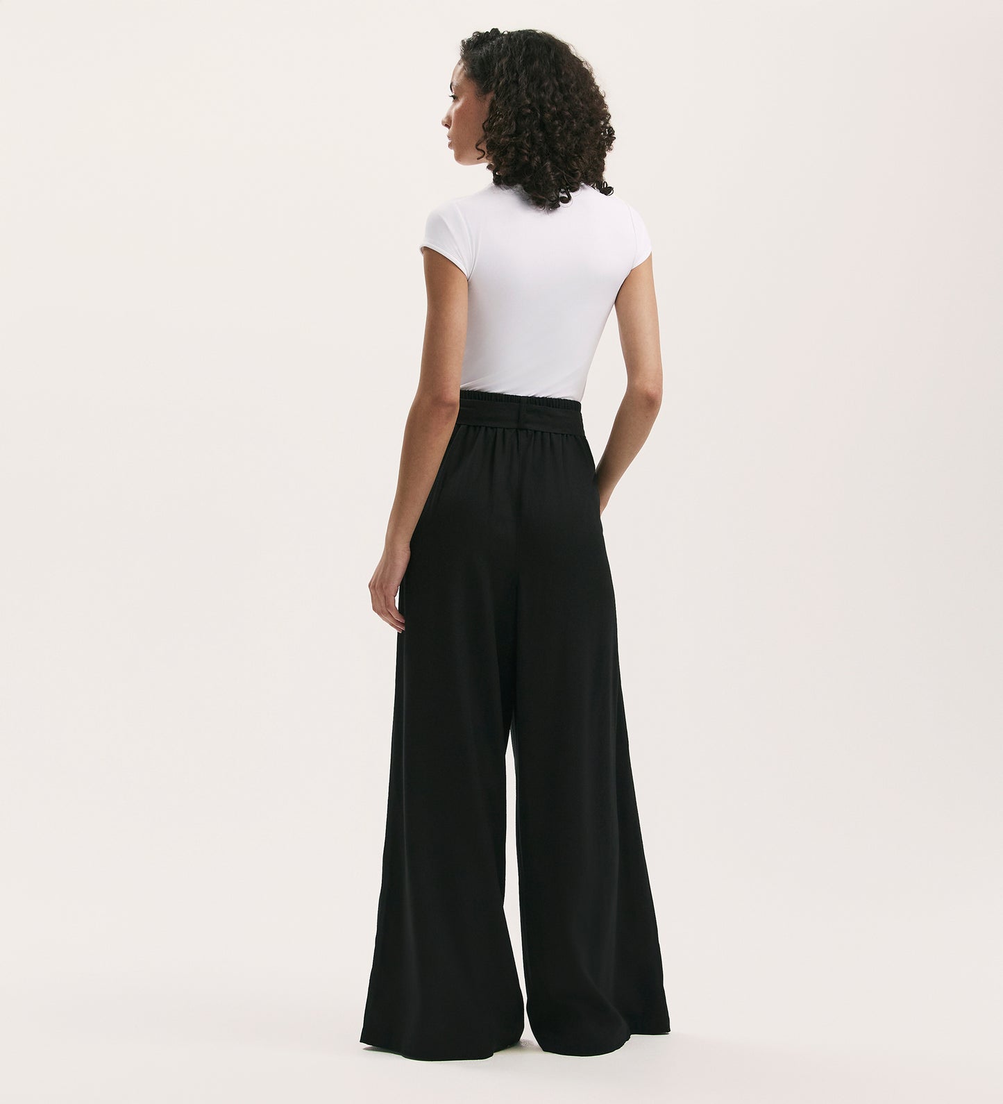 A woman with curly hair stands facing away, wearing a fitted white short-sleeve top and Athena Black Linen Blend Wide Leg Trousers against a plain light background.