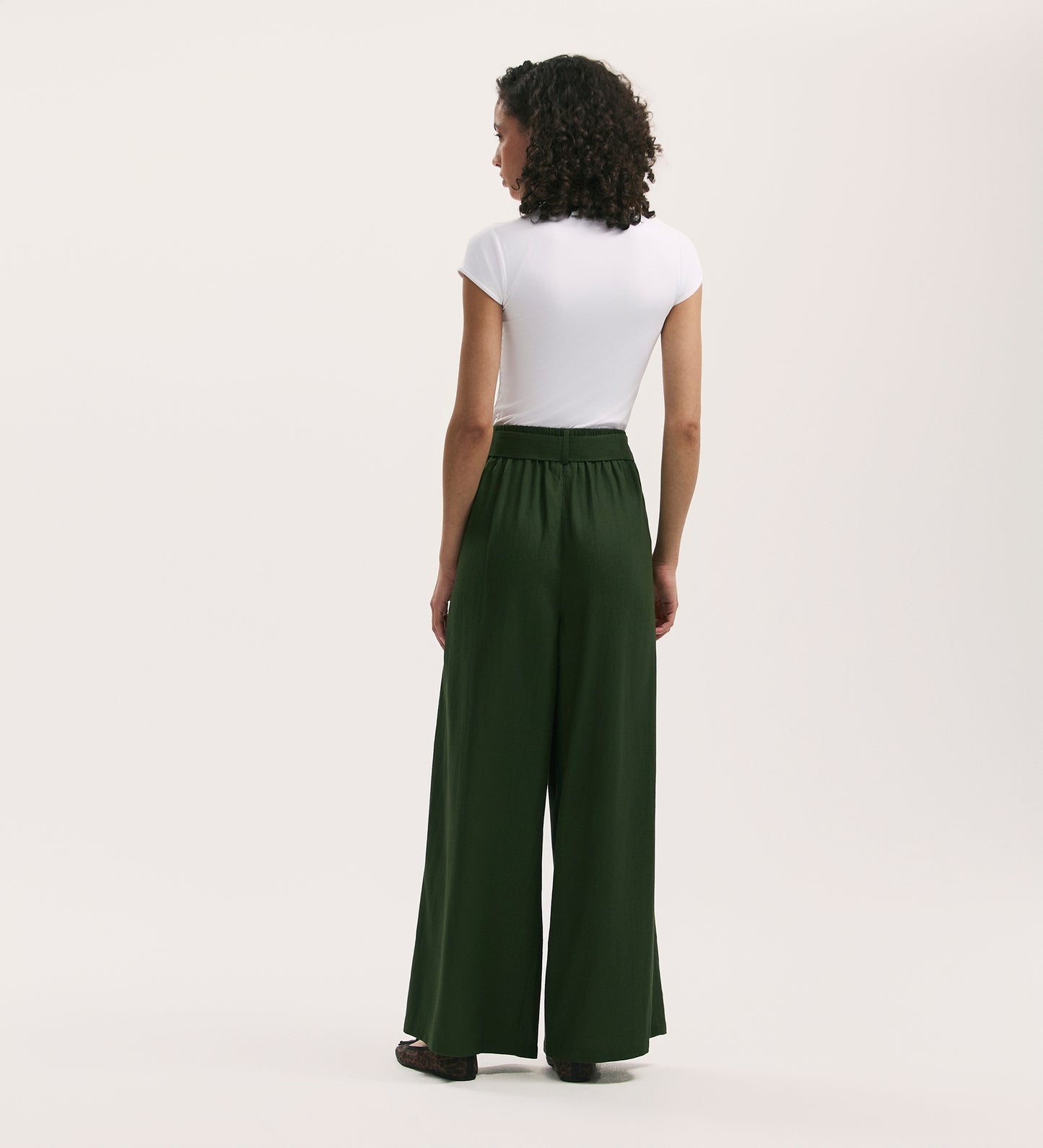 A person with curly hair, wearing a fitted white short-sleeve top and Athena Dark Khaki Linen Mix Wide Leg Trousers, stands facing away against a plain light background.
