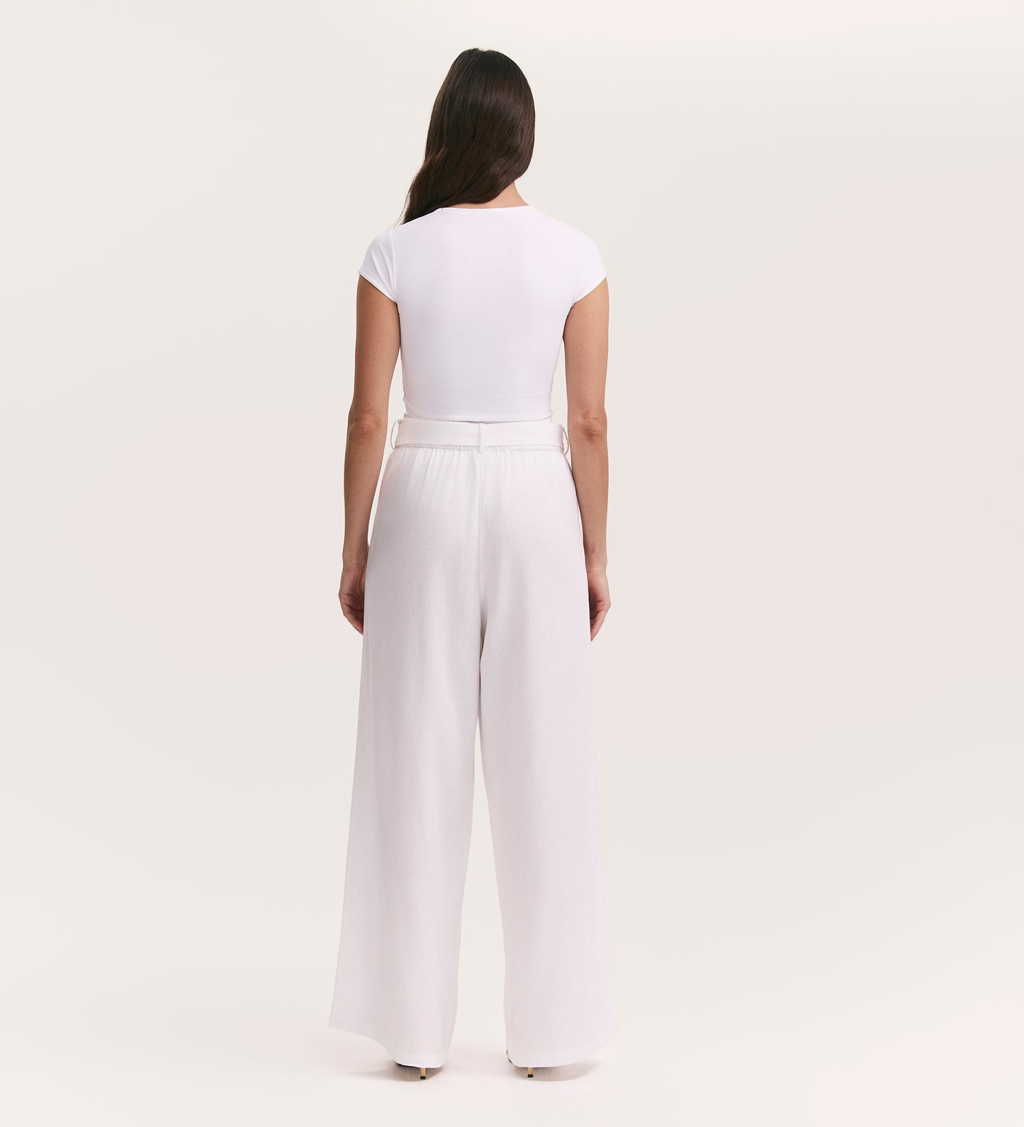 A woman faces away, wearing a fitted white short-sleeve top and Athena White Linen Blend Wide Leg Trousers against a plain light background.