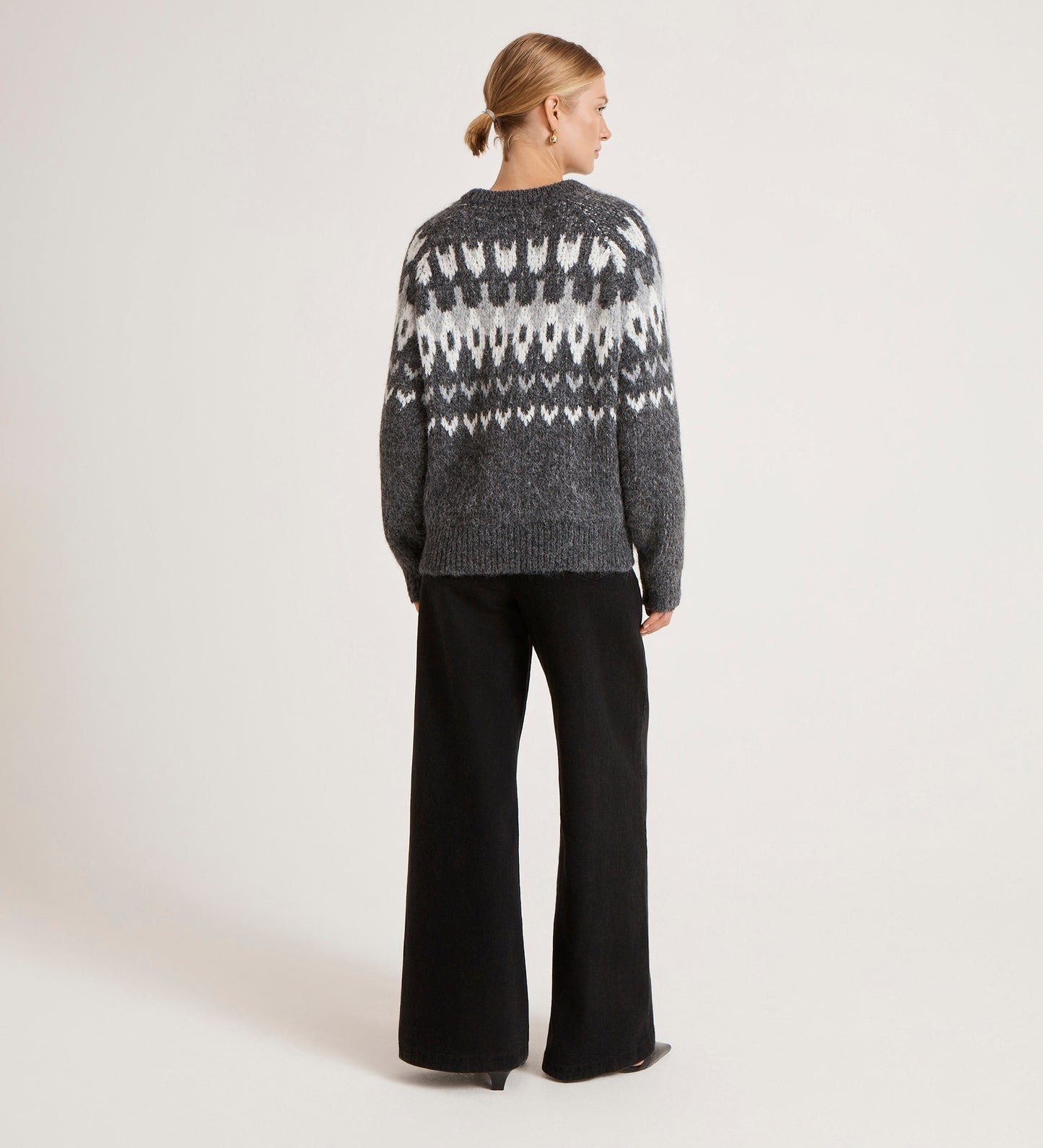 A woman with blonde hair in a low bun stands with her back to the camera, wearing the Belina Grey Marle Knitted Jumper and wide-leg black pants against a plain light background.
