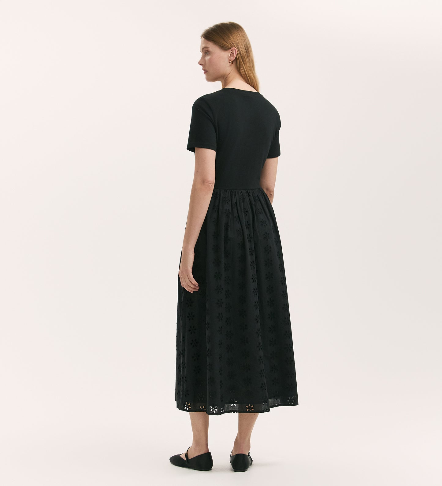 A woman with light brown hair stands facing away, wearing the Brea Black Cotton Midi T-Shirt Dress and black flat shoes. The dress has a relaxed fit and reaches mid-calf. She poses against a plain white background.