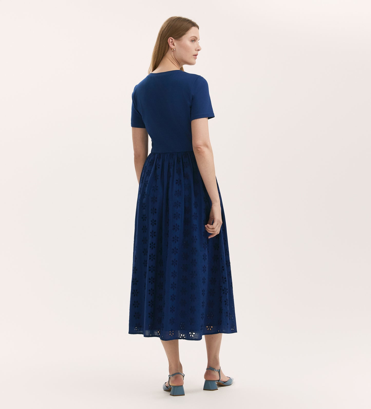 A woman with straight, light brown hair and blue sandals stands against a plain background, wearing the Brea Indigo Cotton Midi T-Shirt Dress with a solid top and an embroidered skirt in soft cotton.