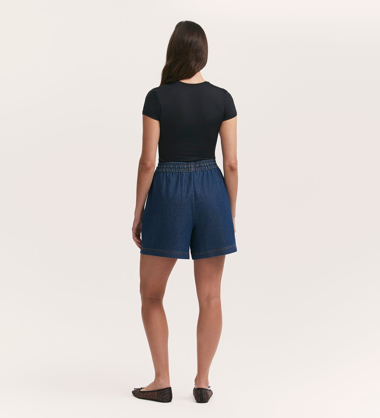 A woman faces away from the camera, wearing a black short-sleeve top and the Britney Indigo Denim Shorts with an elastic waistband and side pockets, paired with black shoes against a plain, light background.