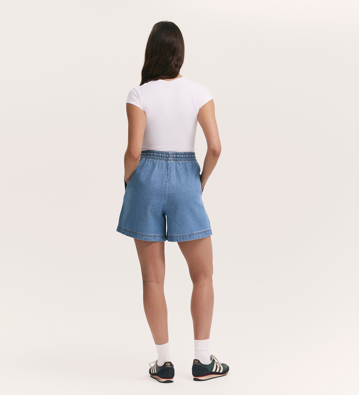 A person with long brown hair, wearing a white t-shirt, Britney Light Blue Denim Shorts with functional side pockets, white socks, and black sneakers stands facing away with hands in their pockets against a plain light background.