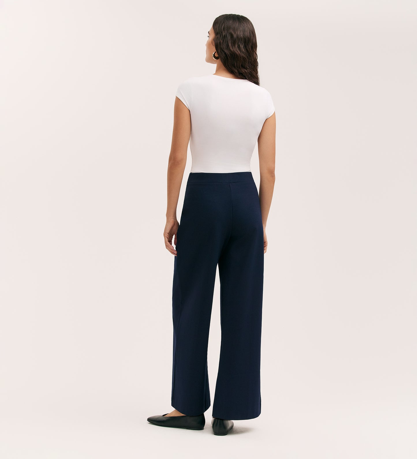 A woman with long wavy hair stands facing away against a plain light background, wearing black flats, a fitted white short-sleeve top, and the Caryl Navy Ponte Jersey Wide Leg Trouser with an elasticated waistband.