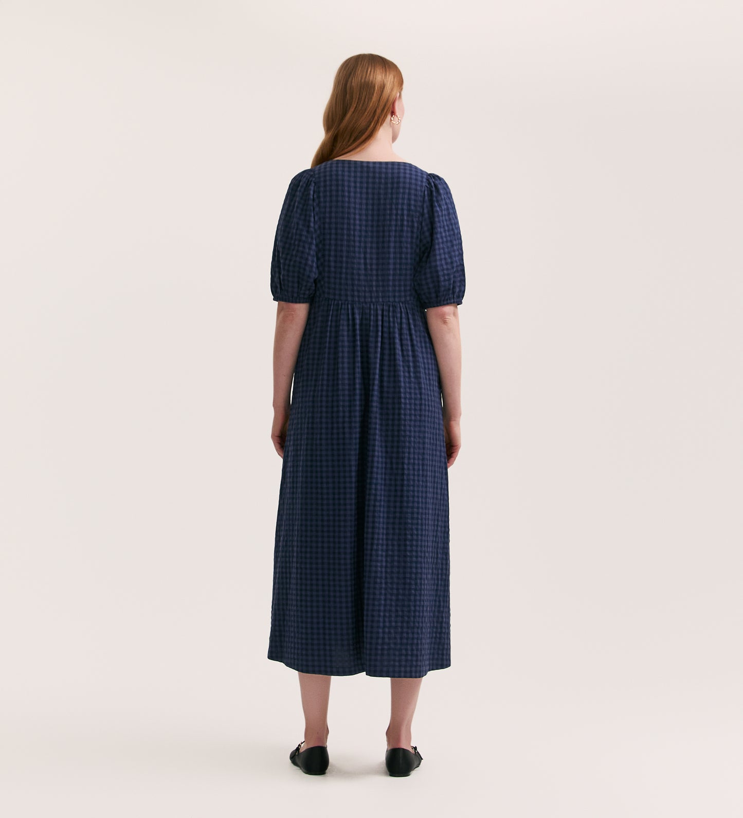 A woman with long red hair stands facing away, wearing the Chloe Navy Gingham Seersucker Midi Dress with short puff sleeves and black shoes. The dress features an oversized fit for effortless style, set against a plain light background.