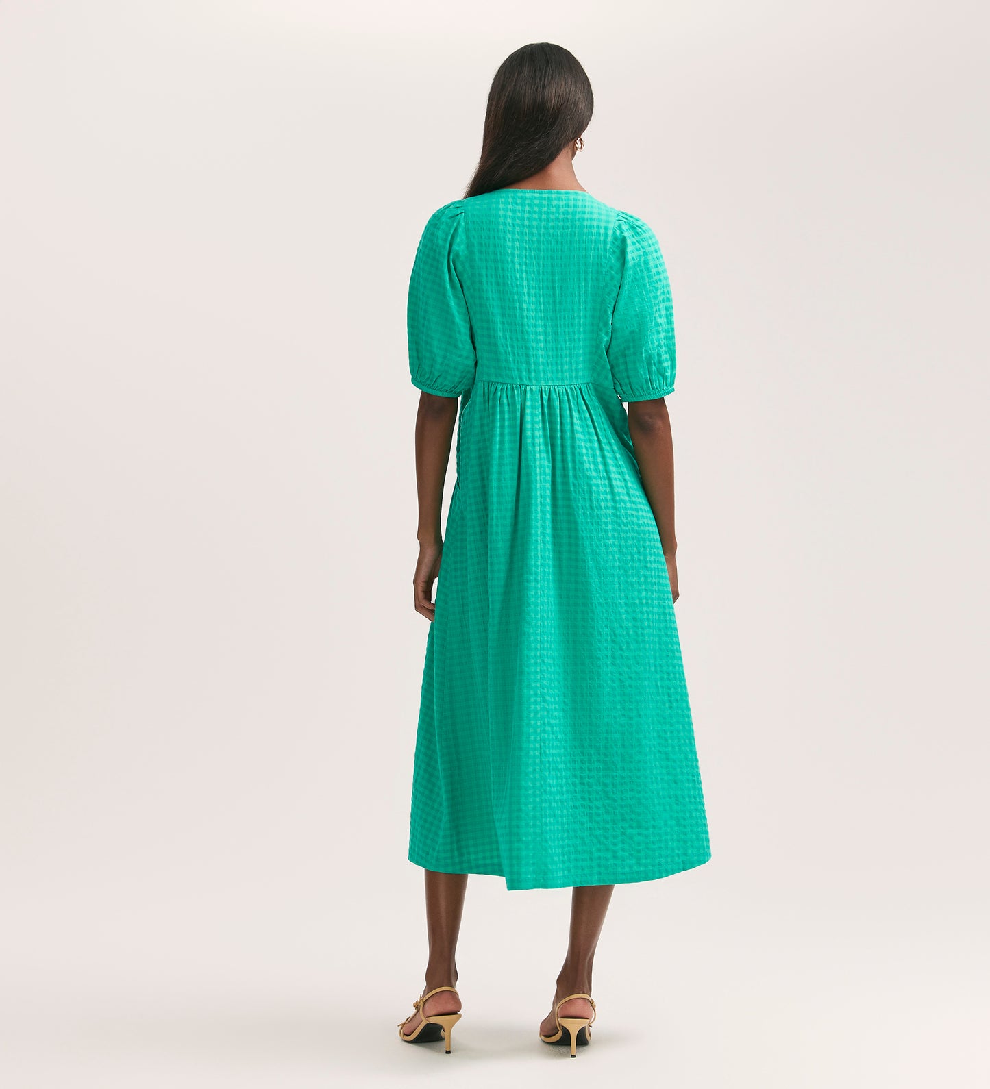 A woman with long hair and heeled sandals faces away, wearing the Chloe Green Gingham Seersucker Midi Dress featuring short puff sleeves and textured cotton seersucker. The background is plain and light-colored.