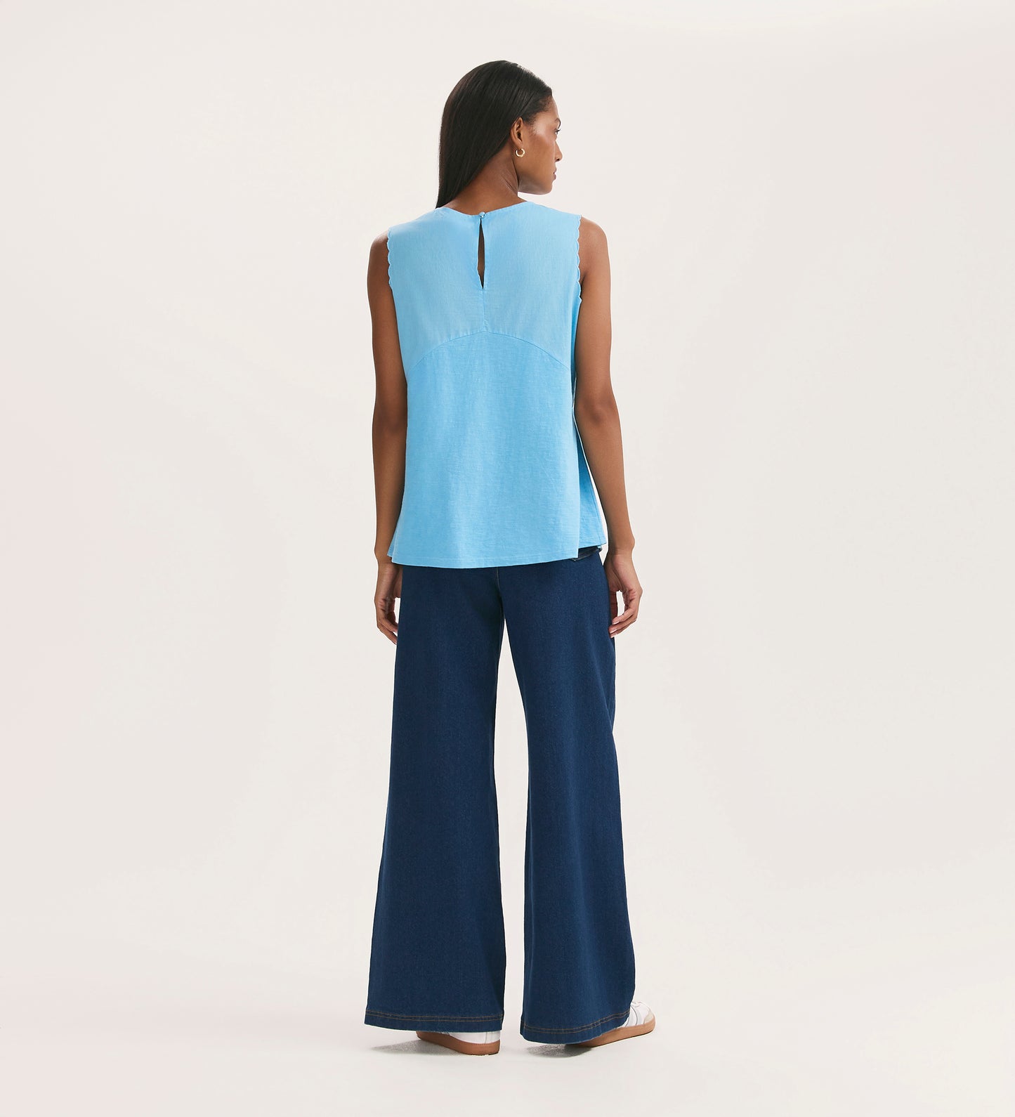 A woman stands facing away, wearing the Chrissy Light Blue Cotton Sleeveless Top and wide-leg dark blue jeans, her long dark hair flowing against a plain light background.