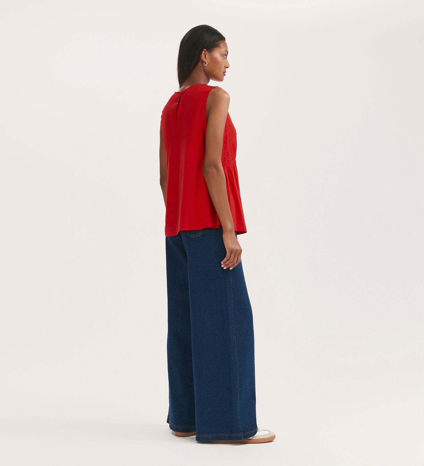 A woman stands sideways against a plain light background wearing the Chrissy Red Cotton Sleeveless Top, featuring scallop detailing, wide-leg blue jeans, and white shoes.