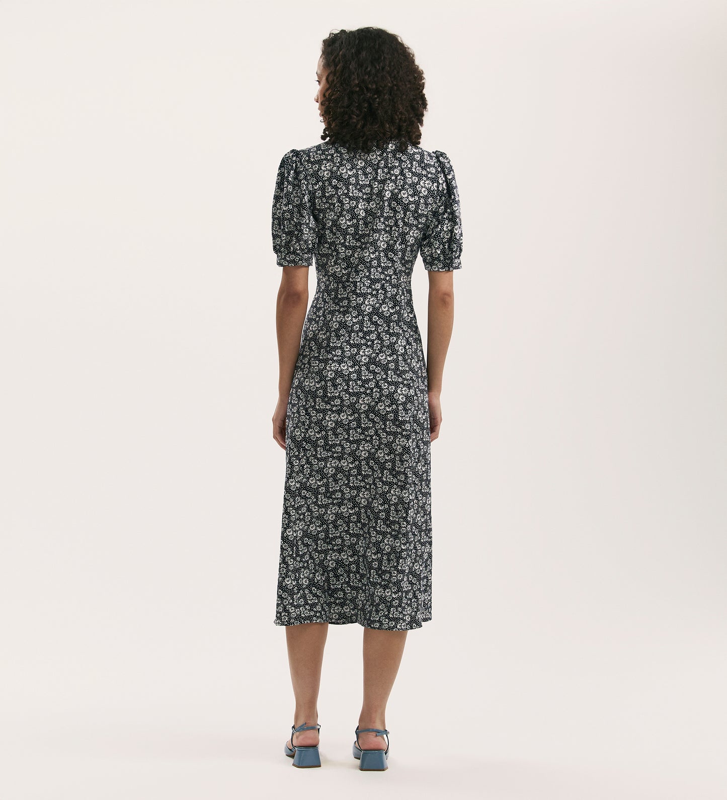 A person with curly hair wears the Colette Navy Daisy Crepe Midi Tea Dress—featuring a white floral pattern and short puff sleeves—paired with blue heeled sandals, facing away from the camera against a light background.
