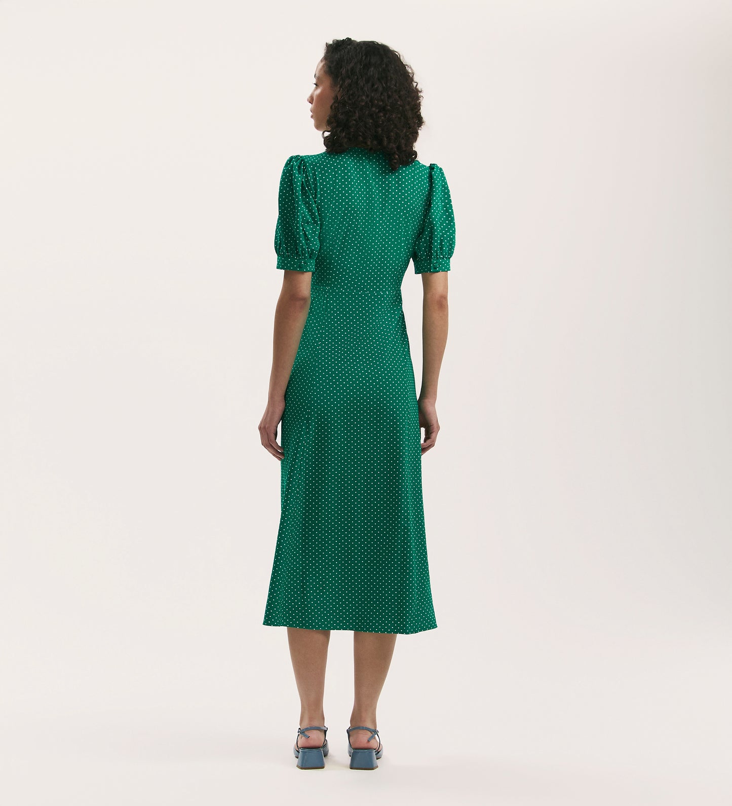 A person with curly hair faces away, wearing the Colette Green Polka Dot Crepe Midi Tea Dress with puffed shoulders and blue heels. The crease-free crepe fabric adds elegance against a plain light background.