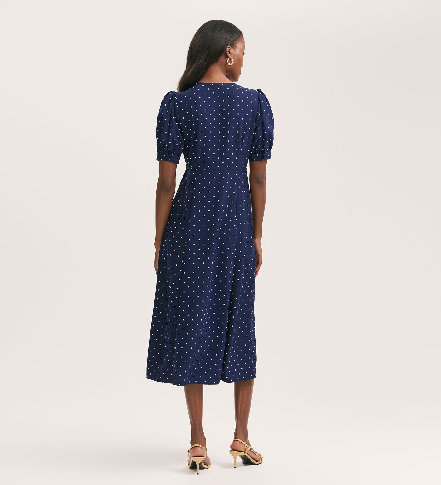 A woman with long hair wears the Colette Navy Polka Dot Crepe Midi Tea Dress, featuring puffed sleeves and an empire waist. The crease-free crepe fabric dress is paired with heeled sandals against a plain, light background.