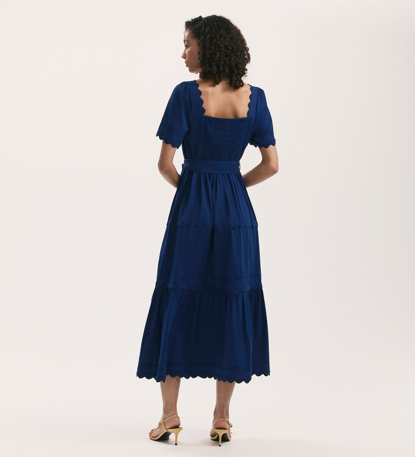 A woman with curly hair stands facing away, modeling the Corina Indigo Cotton Midi Tiered Dress with a square neck and scalloped hem, paired with yellow heeled sandals against a plain light background.