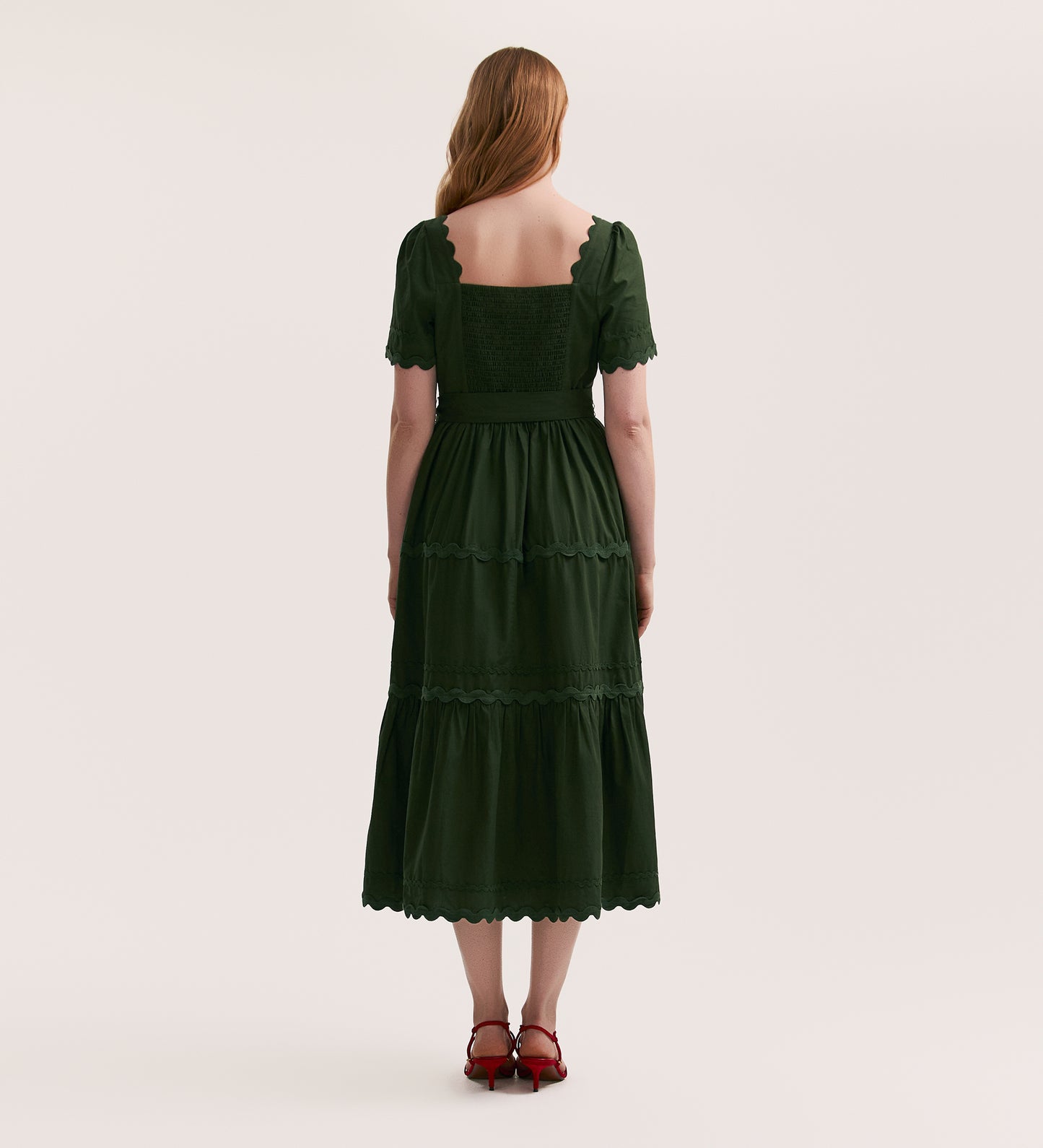 A woman with long, wavy hair stands facing away in the Corina Dark Khaki Cotton Midi Dress, featuring scalloped edges and a tiered skirt, paired with red heeled sandals against a plain light background.