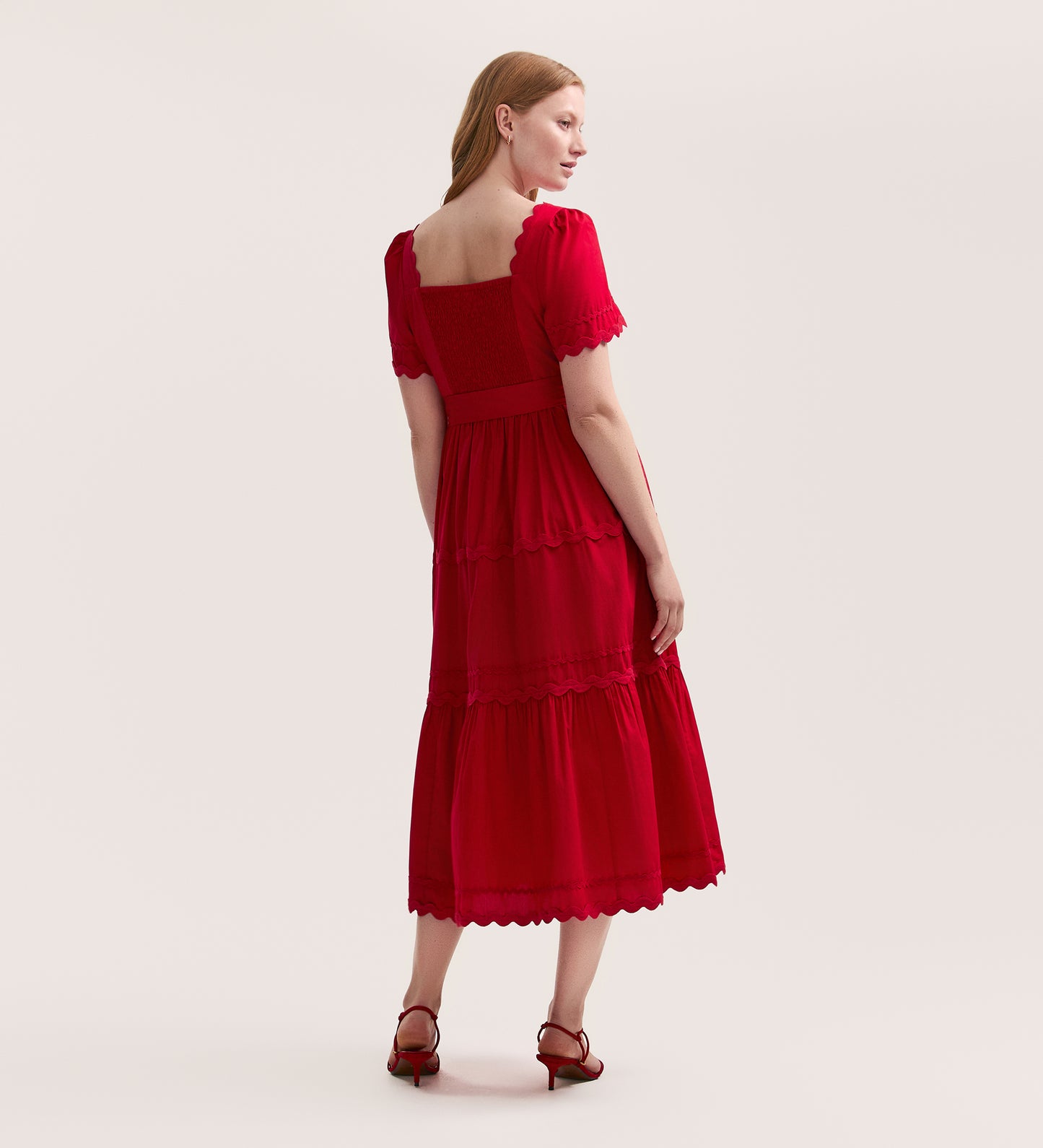 A woman with light skin and long blond hair stands with her back to the camera, wearing the Corina Red Cotton Midi Dress featuring scalloped edges and tiered layers, paired with red heeled sandals.