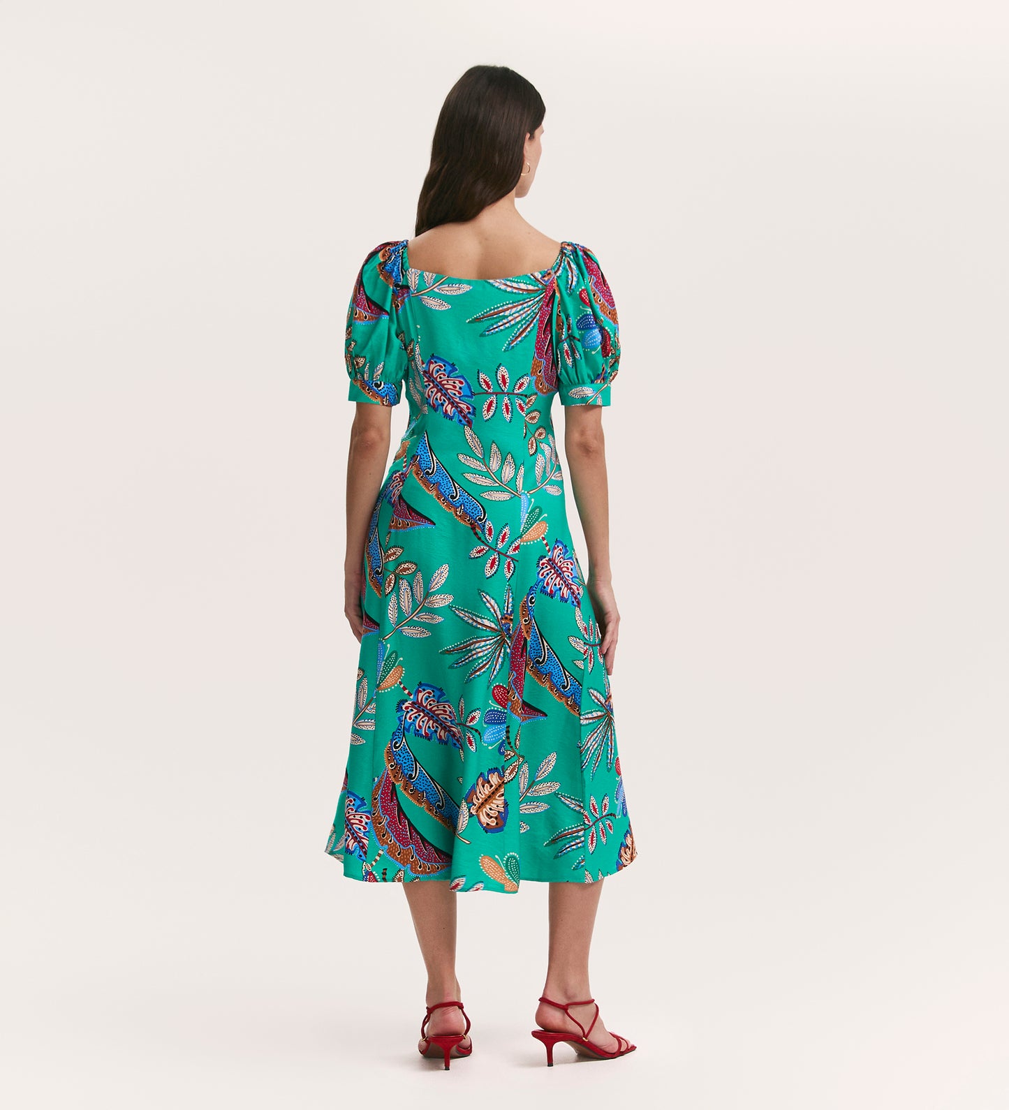 A woman with long brown hair faces away, wearing the Daisy Green Leaves Crepe Midi Dress—a flattering, fit-and-flare teal midi with colorful tropical floral and bird patterns, puffed short sleeves, and paired with red strappy heels.
