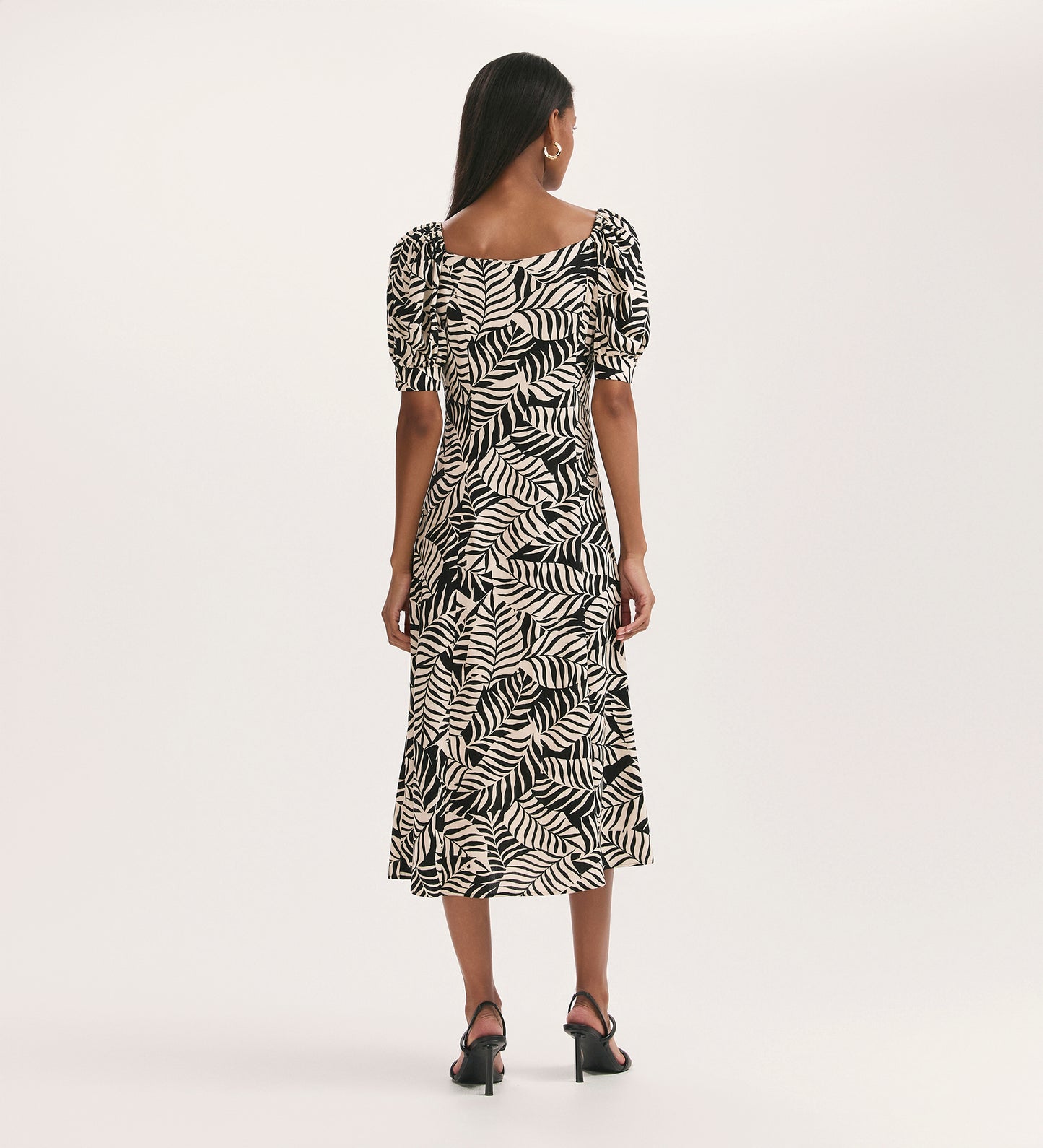 A woman faces away, wearing the Daisy Black Leaves Crepe Midi Dress with short puff sleeves, square neck, and fit-and-flare silhouette, paired with black heeled sandals against a plain light background.