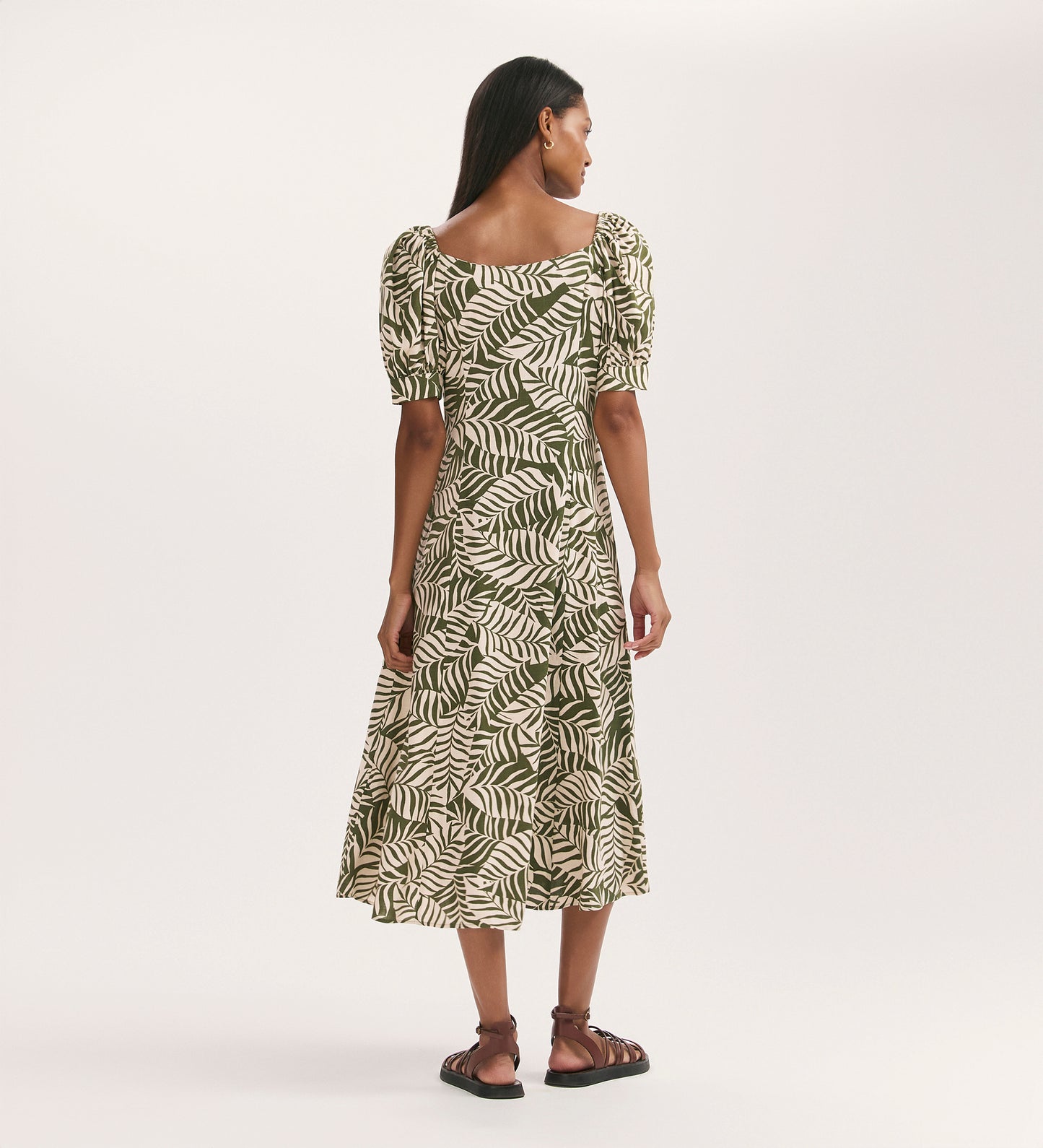 A woman faces away, wearing the Daisy Khaki Leaves Crepe Midi Dress—a cream fit-and-flare midi with green leaf print and puffed short sleeves. She pairs it with brown sandals against a plain light background.