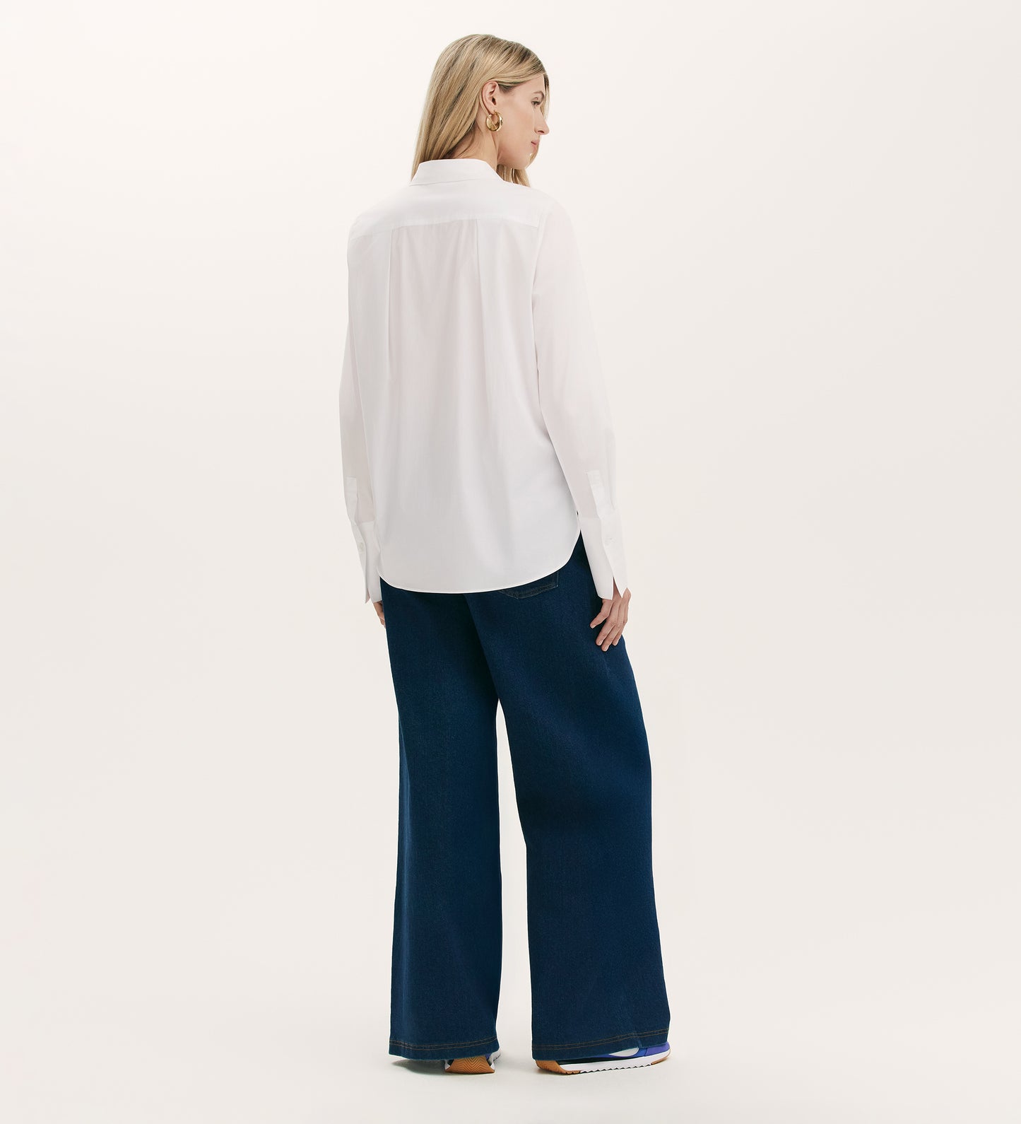 A woman with straight, shoulder-length blonde hair stands facing away against a plain light background, wearing the Dillon White Cotton Shirt paired with wide-leg dark blue jeans.