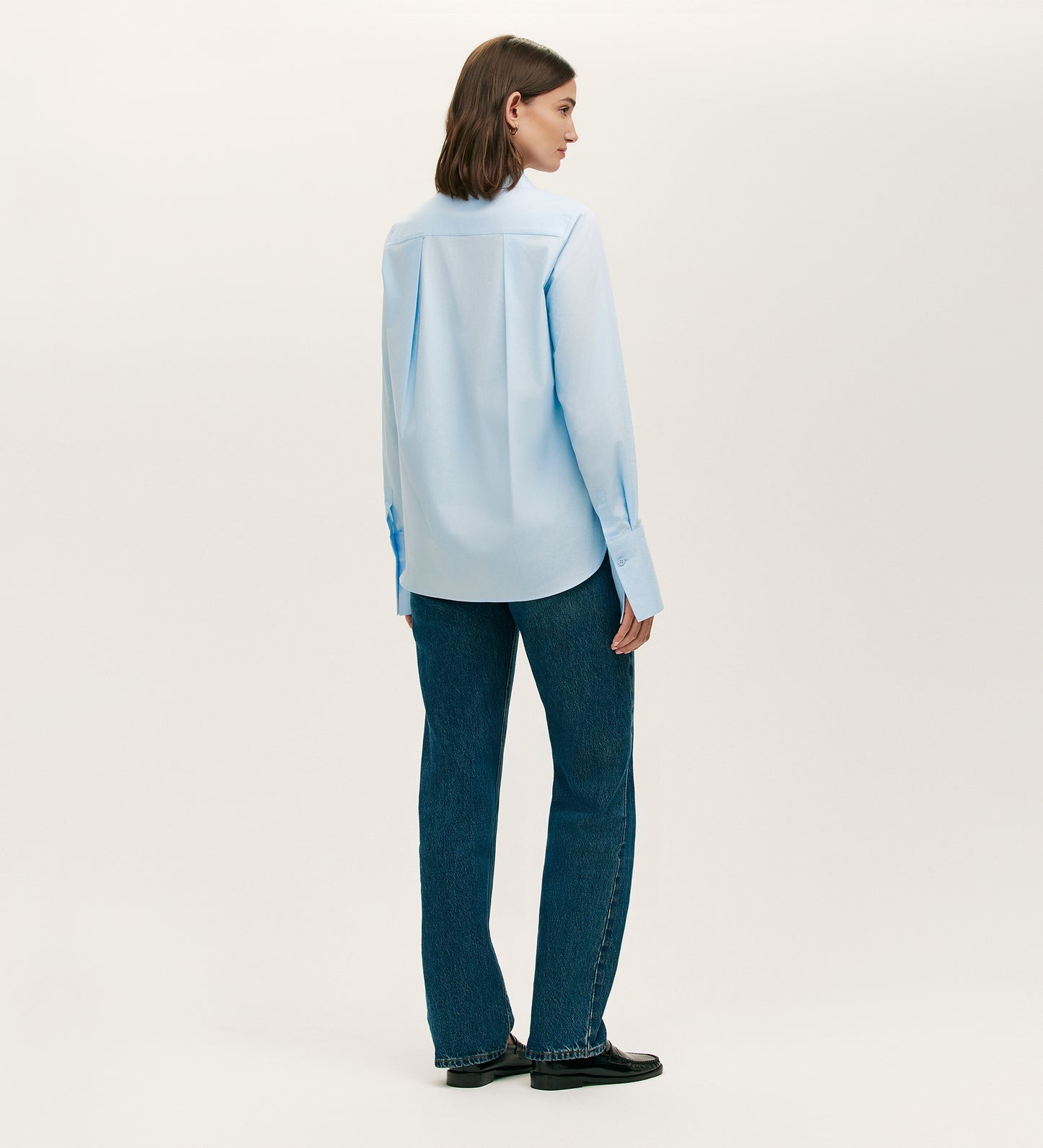 A woman with straight brown hair faces away, wearing the Dillon Blue Cotton Shirt, dark blue jeans, and black shoes against a plain white background.