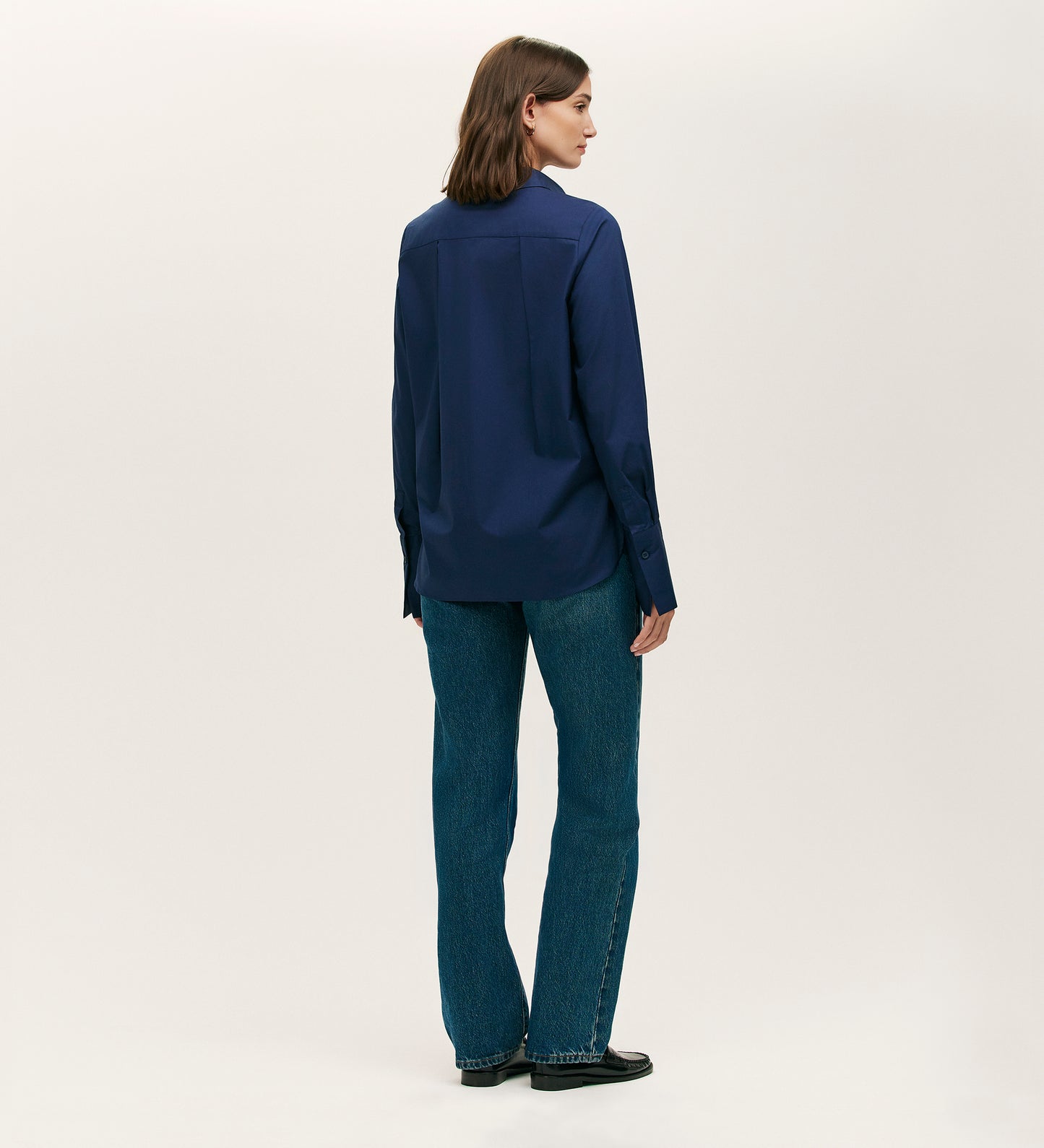 A person with shoulder-length brown hair faces away, wearing the Dillon Navy Cotton Shirt, blue jeans, and black shoes against a plain light background.