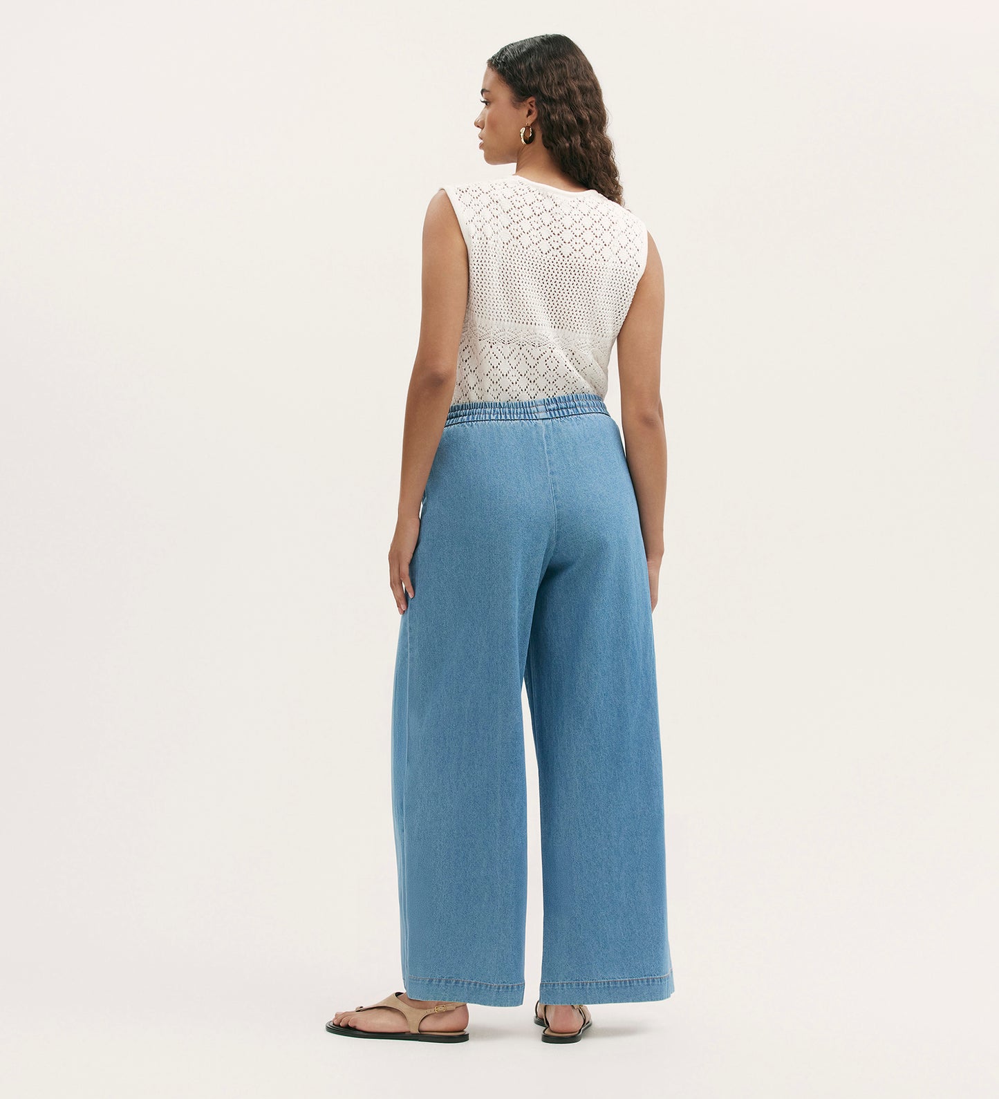 A woman stands facing away, wearing Dixie Light Blue Denim Wide Leg Trousers with an elasticated waistband, styled with a sleeveless white crochet top, black sandals, hoop earrings, and her long wavy hair flowing against a plain light background.