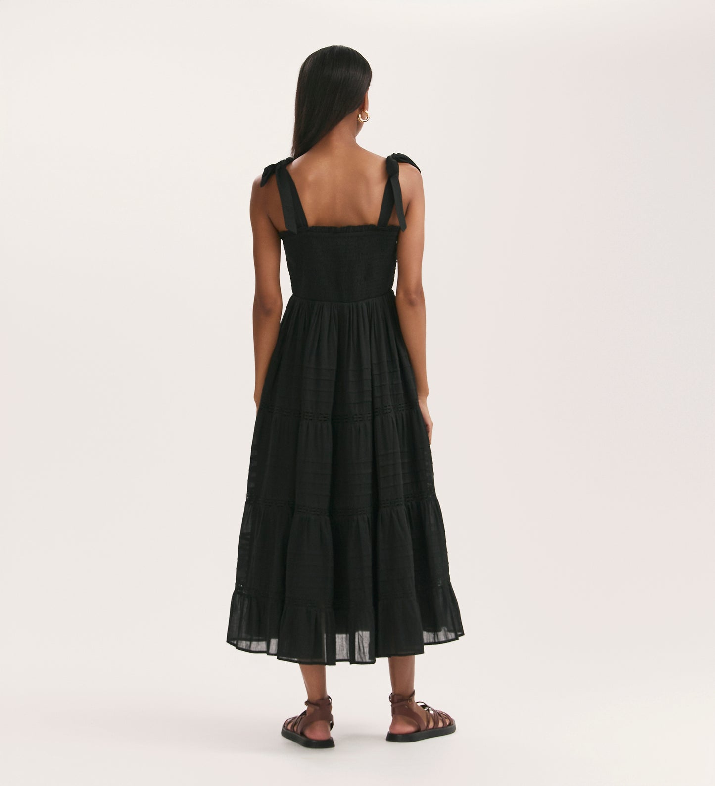 A woman with long dark hair faces away, wearing the Ebony Black Cotton Midi Tiered Dress with tied shoulder straps and a shirred bodice, paired with black flat sandals, set against a plain light background.