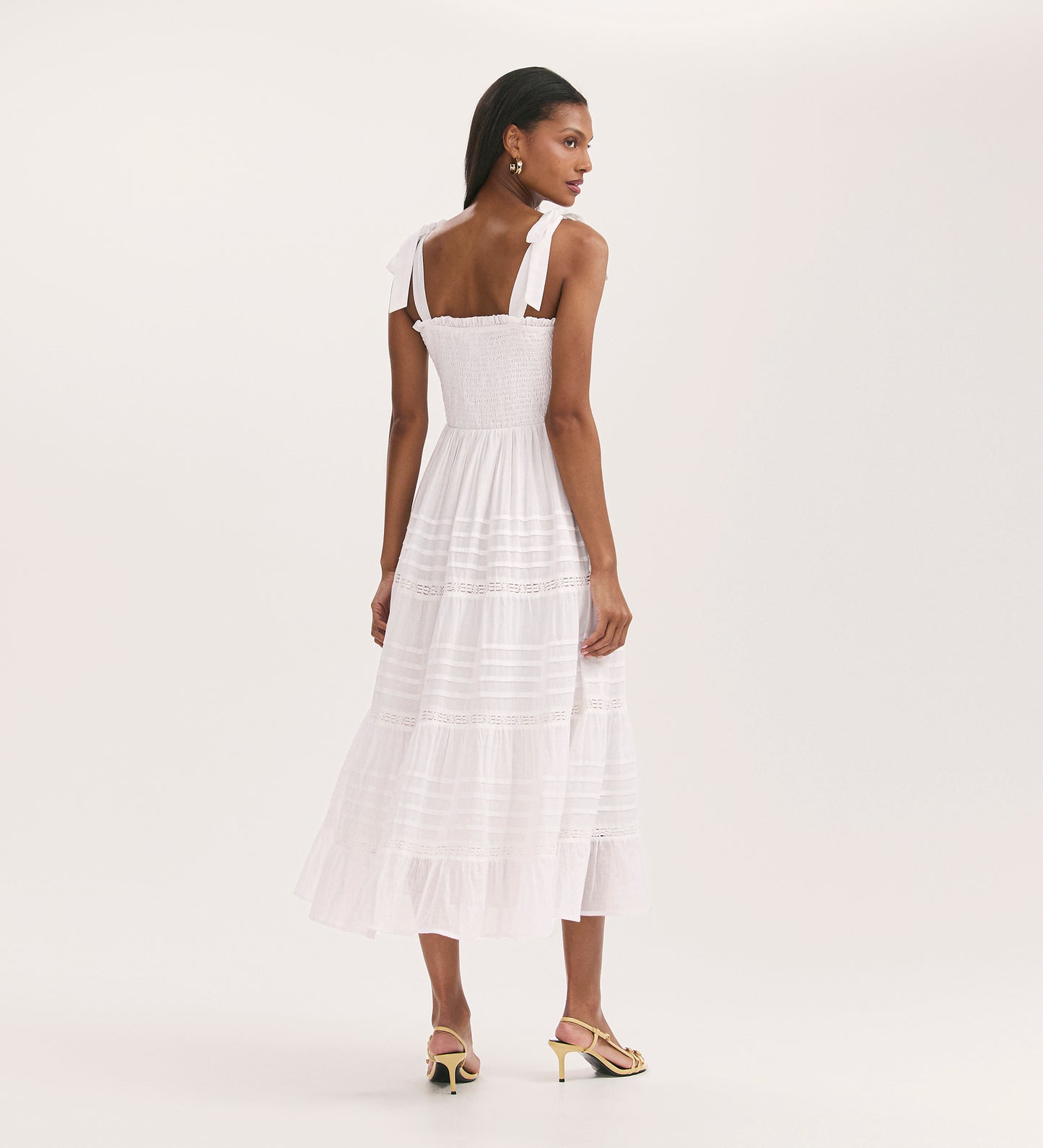 Woman with straight dark hair stands facing away in the Ebony White Cotton Midi Tiered Dress featuring a shirred bodice, shoulder ties, lace details, and open-toe heels against a plain white background.