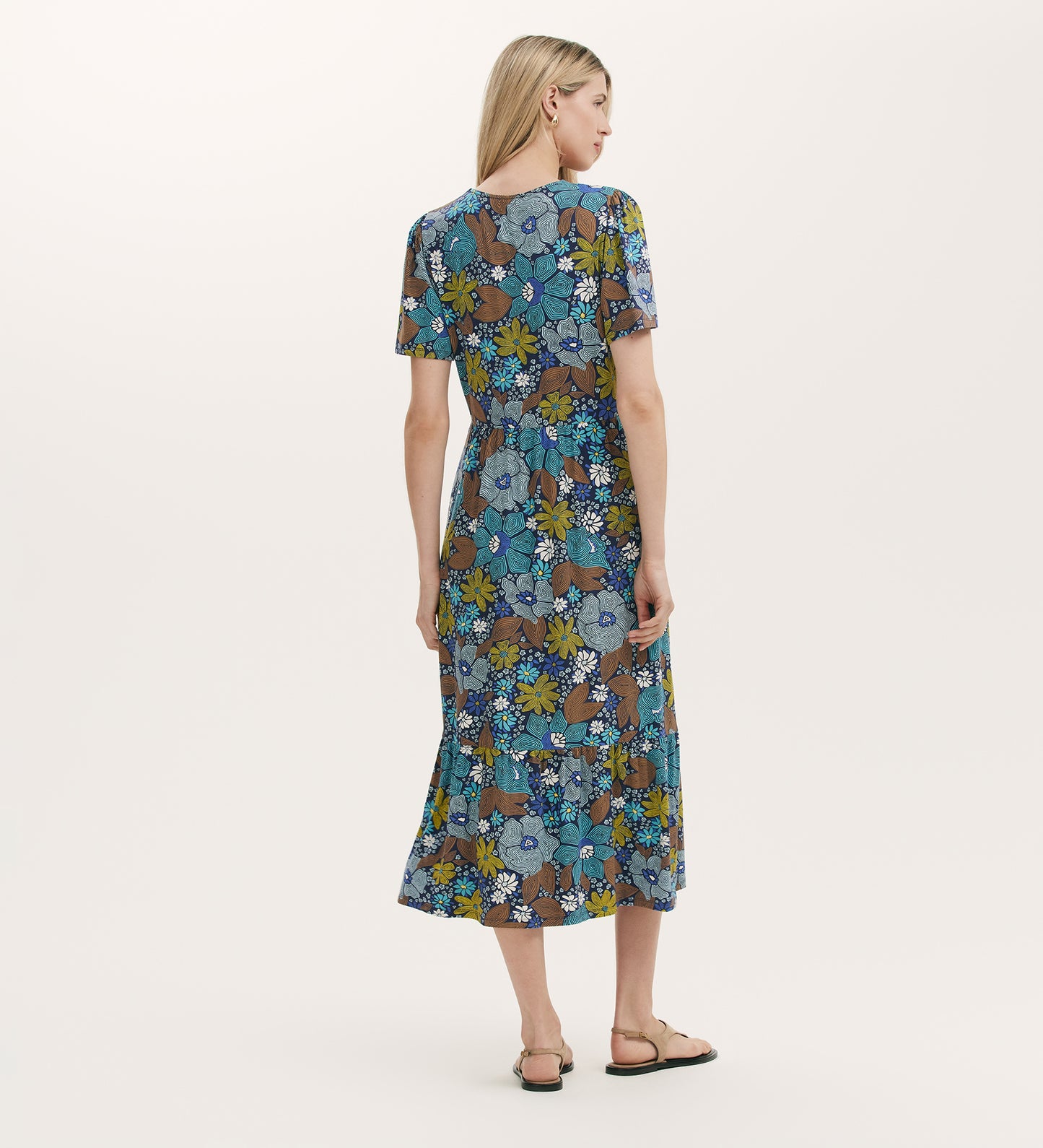 A woman with long blonde hair models the Edith Blue Floral Jersey Midi Dress, featuring a relaxed fit and floral print. She faces away, wearing flat sandals against a plain light background.