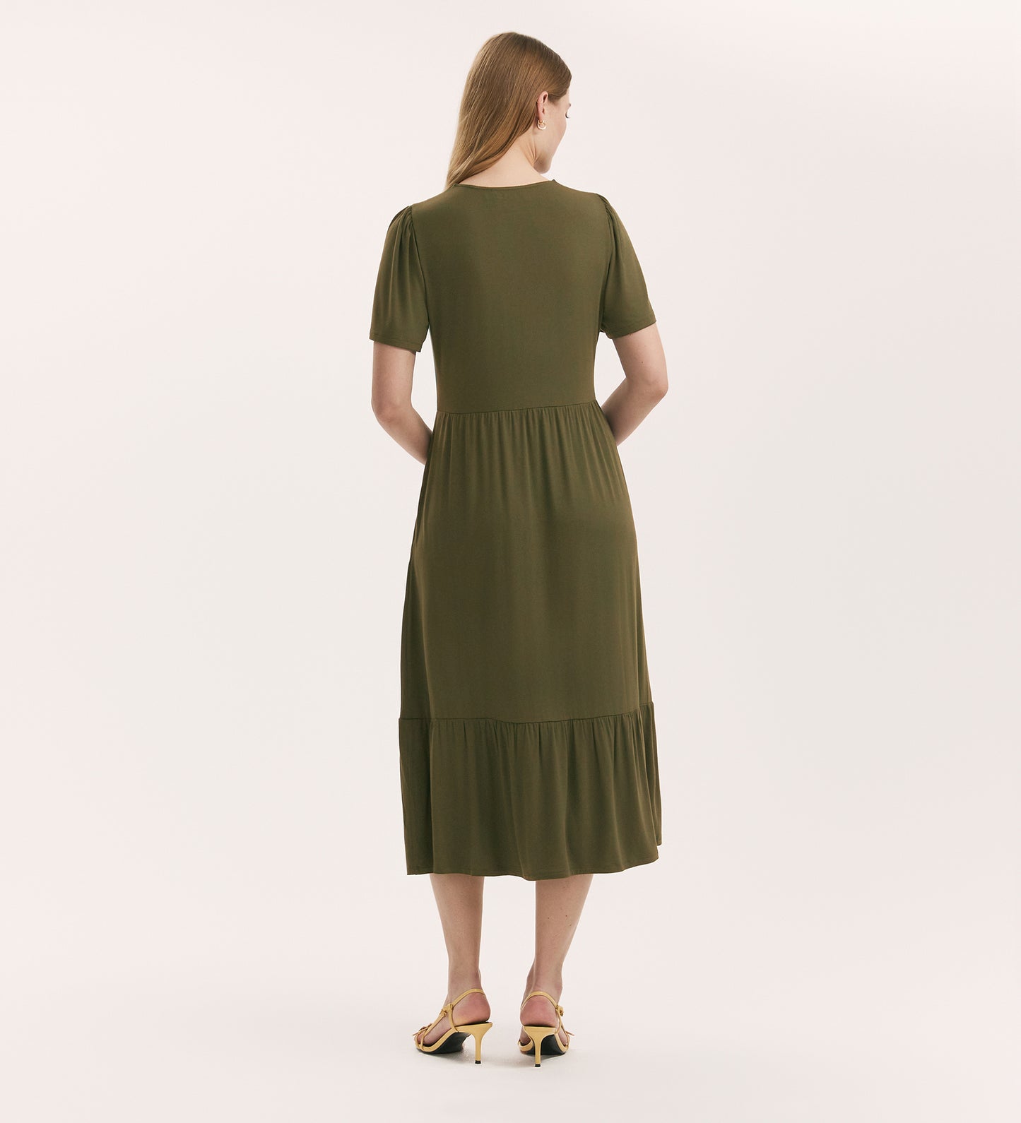 A woman with long blonde hair stands with her back to the camera, wearing the Edith Khaki Jersey Midi Dress and yellow heeled sandals against a plain light background.