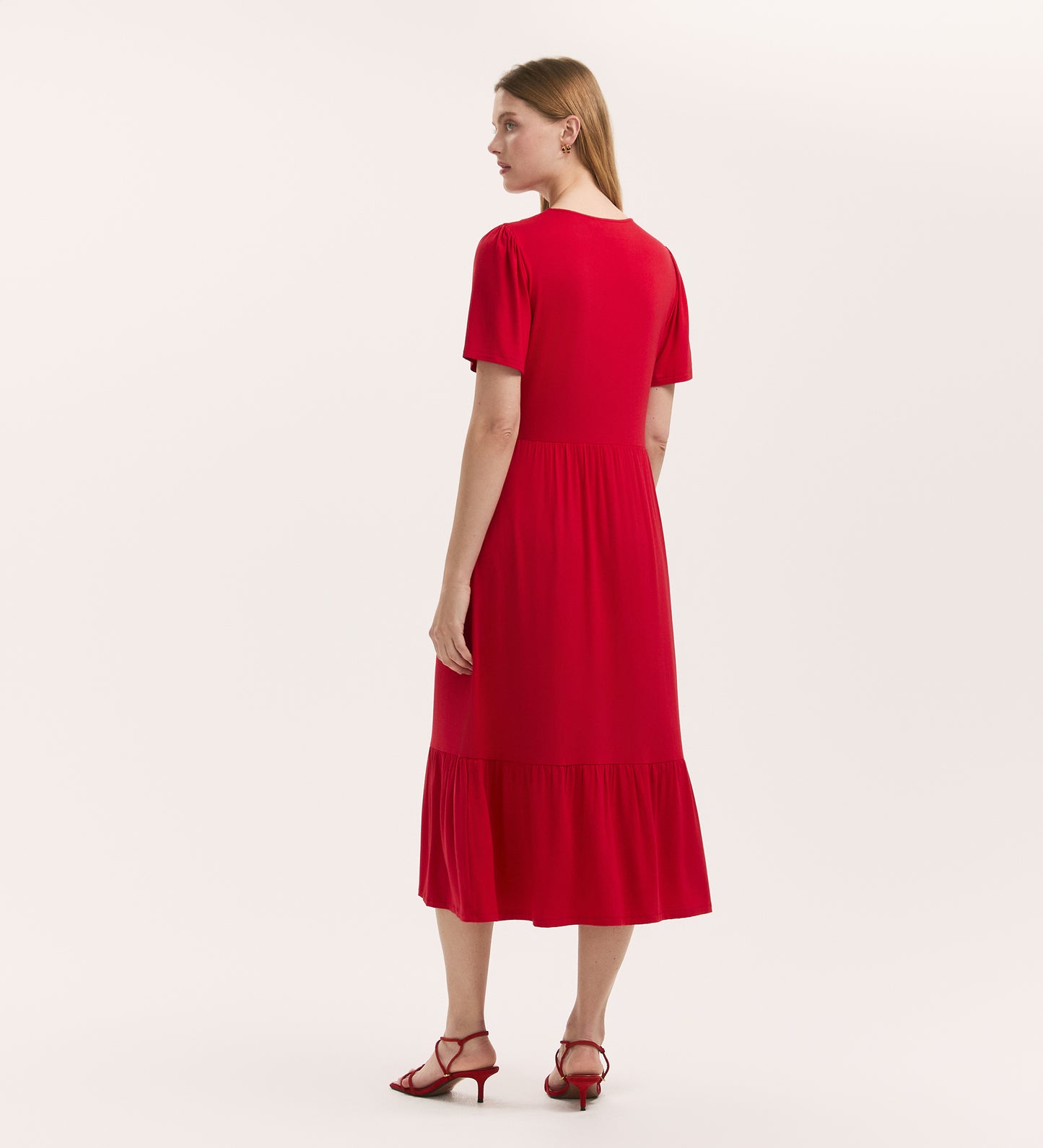 A woman faces away against a white background, wearing the Edith Red Jersey Midi Dress—featuring short sleeves, a button front, gathered skirt—and red heeled sandals.