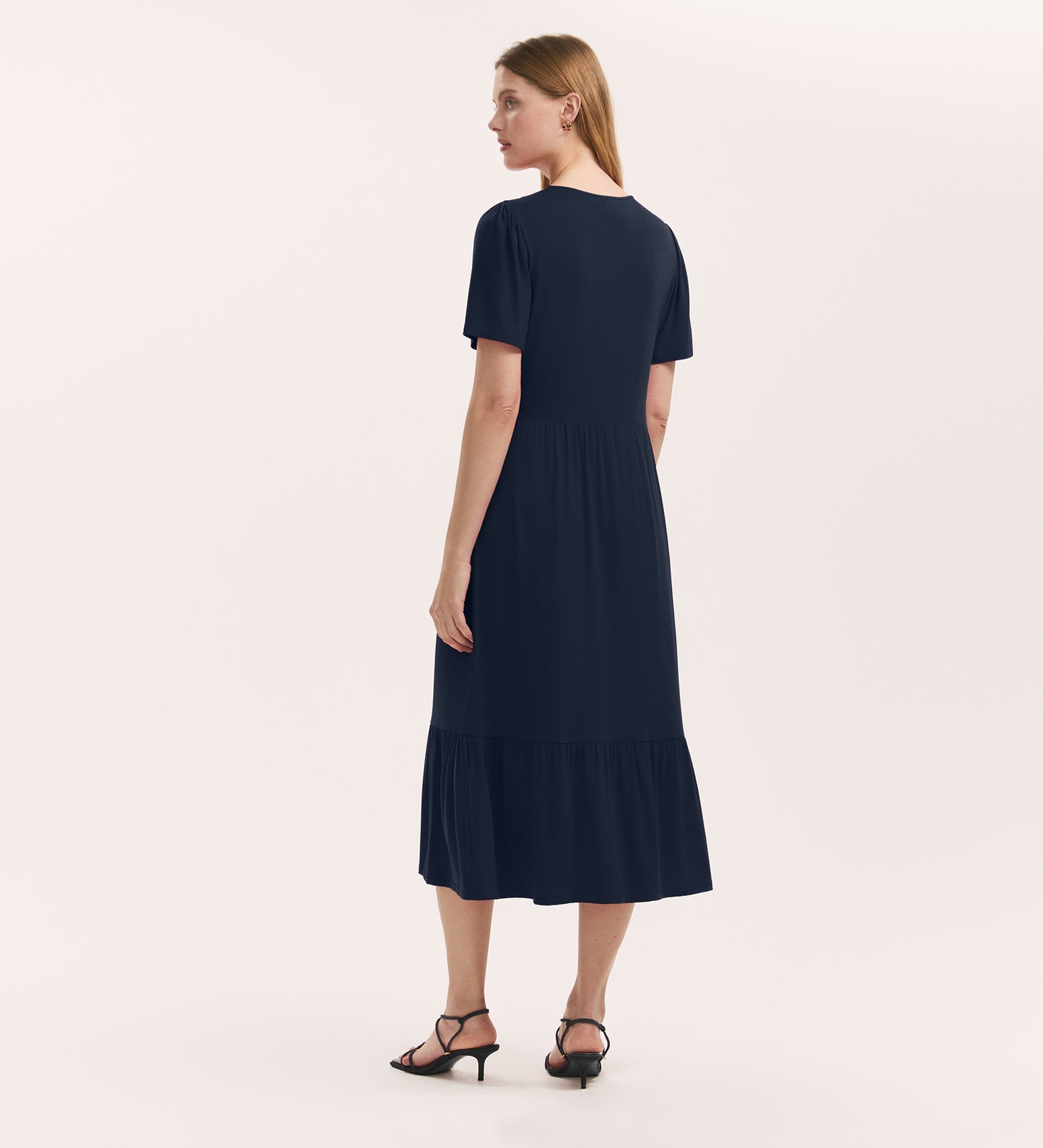A woman with straight, light brown hair stands facing away, wearing the Edith Navy Jersey Midi Dress with a v-neck and flared hem, paired with black heeled sandals against a white background.