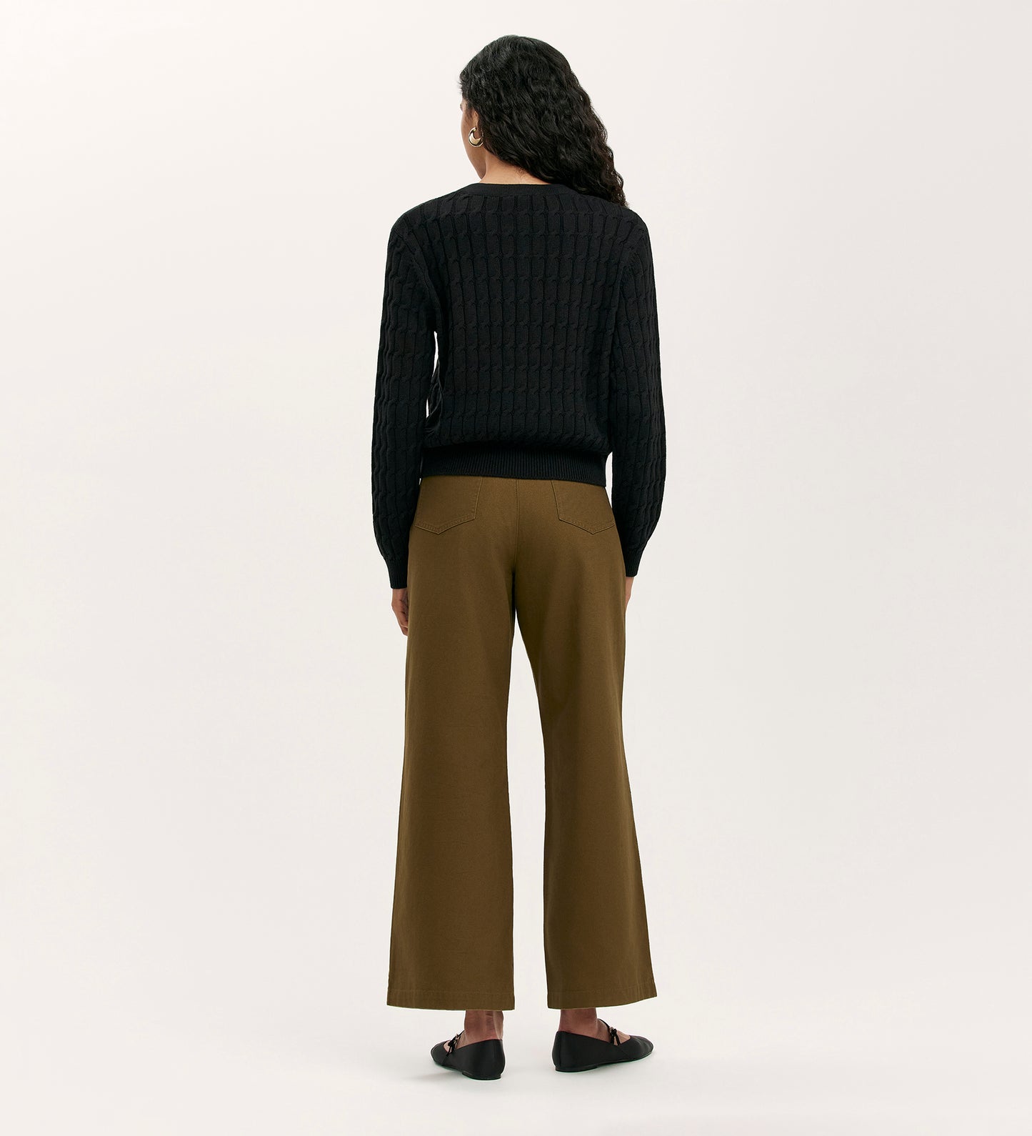 A person with long wavy hair stands facing away, wearing the Elaine Black Knitted Cardigan, brown wide-leg pants, and black flats against a plain light background.