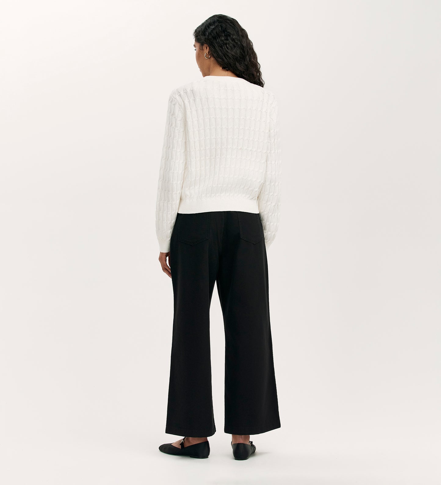 A person with long dark hair, wearing the Elaine Ivory Knitted Cardigan paired with black wide-leg pants and black shoes, stands facing away against a plain white background.