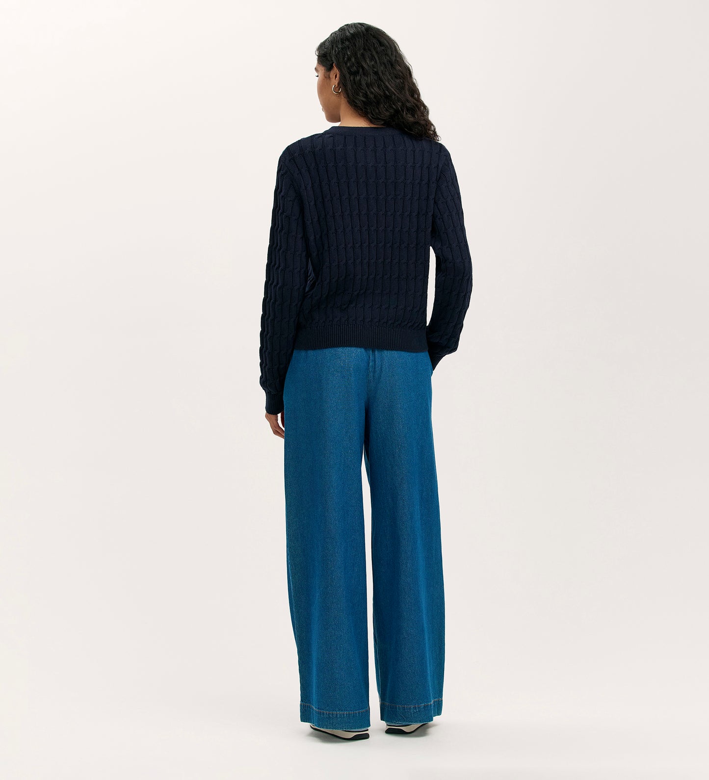A person with long dark hair faces away, wearing the Elaine Navy Knitted Cardigan with gold buttons and wide-leg blue jeans, set against a plain, light-colored background.