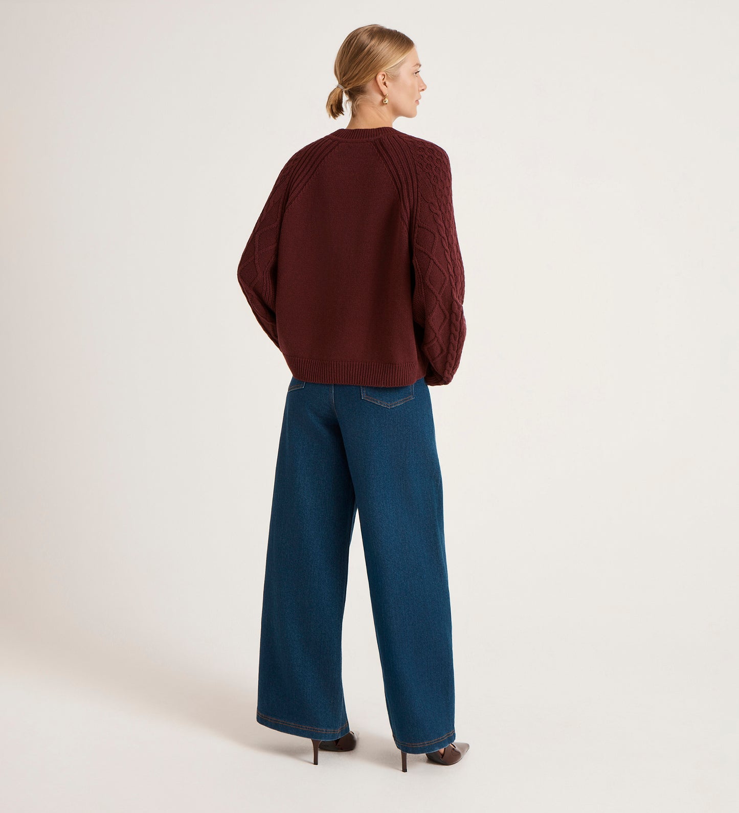 A person with blonde hair in a low bun, wearing the Ellie Burgundy Knitted Jumper, wide-leg blue jeans, and dark heels, stands facing away in a plain light-colored studio.