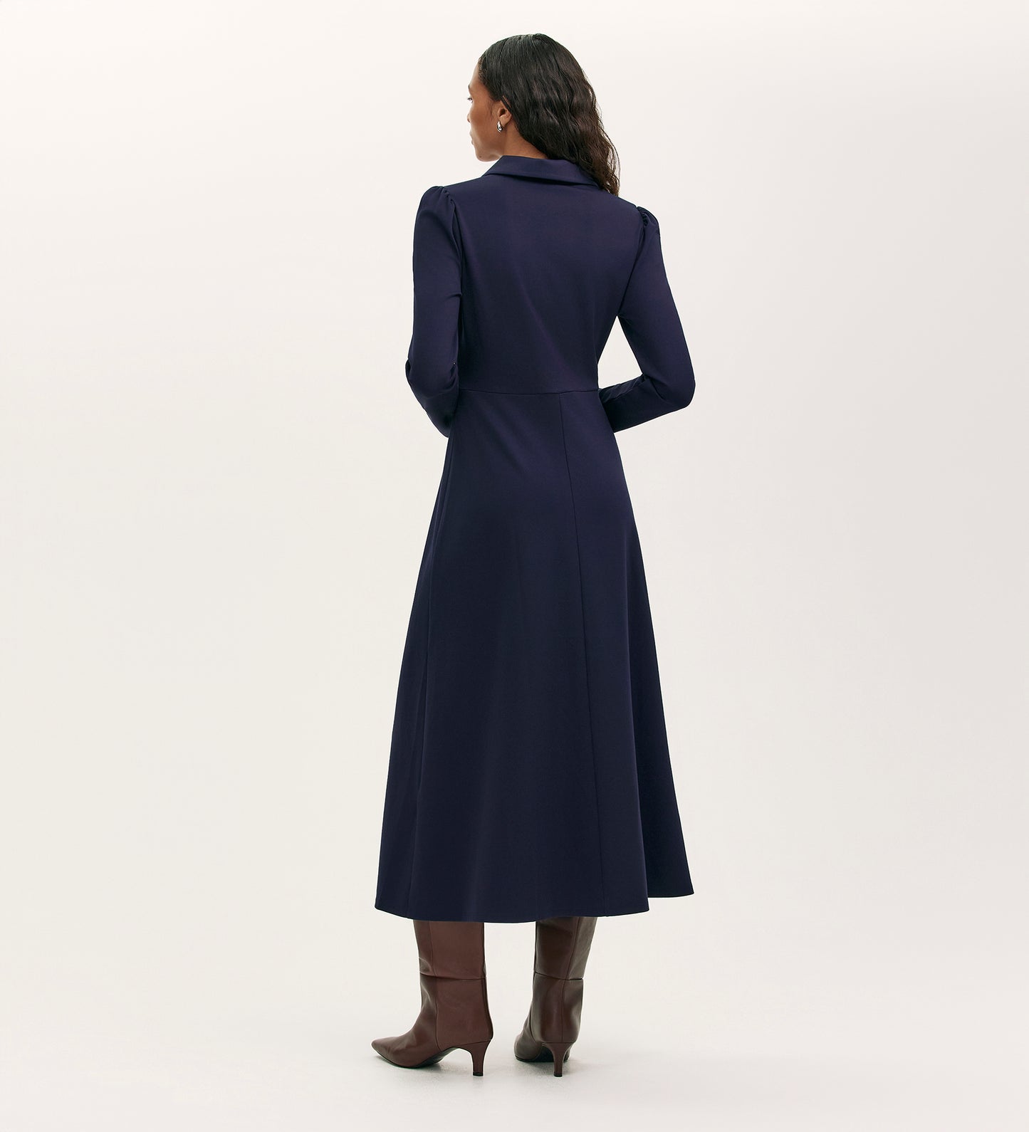 A woman with wavy hair faces away, wearing the Elvira Navy Jersey Midi Dress featuring a collared neckline and long sleeves. She pairs it with brown heeled boots, standing against a plain light background.