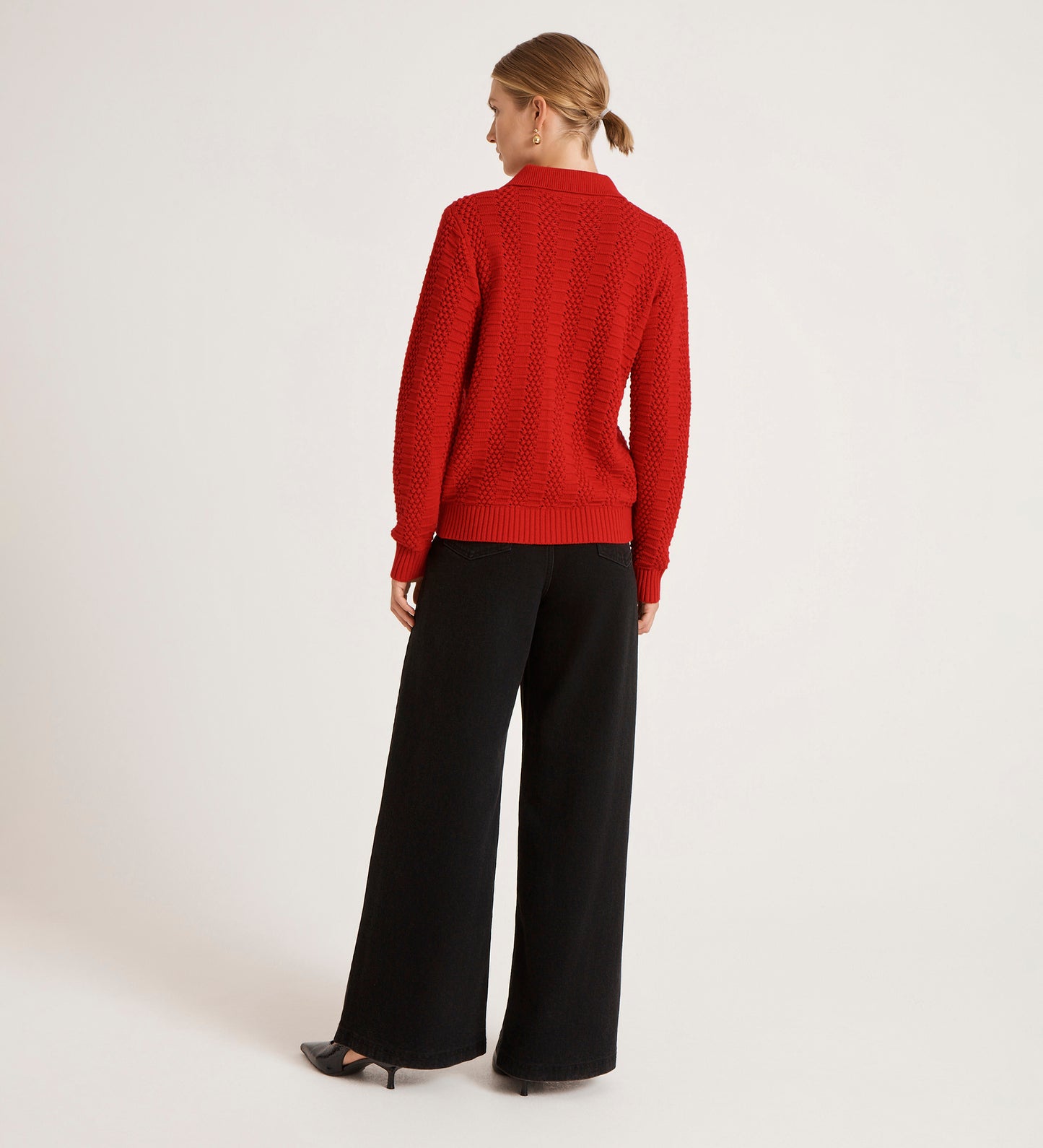 A woman stands facing away, wearing the Emlyn Red Knitted Cardigan with wide-leg black pants and black heels, set against a plain white background.