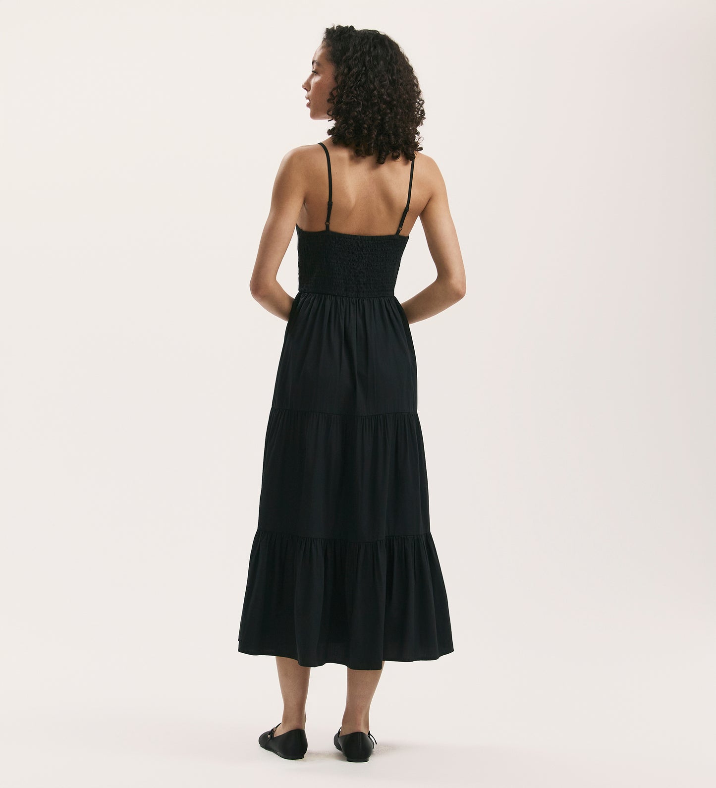 A woman with curly hair stands facing away, wearing the Emmie Black Cotton Midi Dress featuring strappy details and a tiered cotton poplin skirt, paired with black flats against a plain light background.