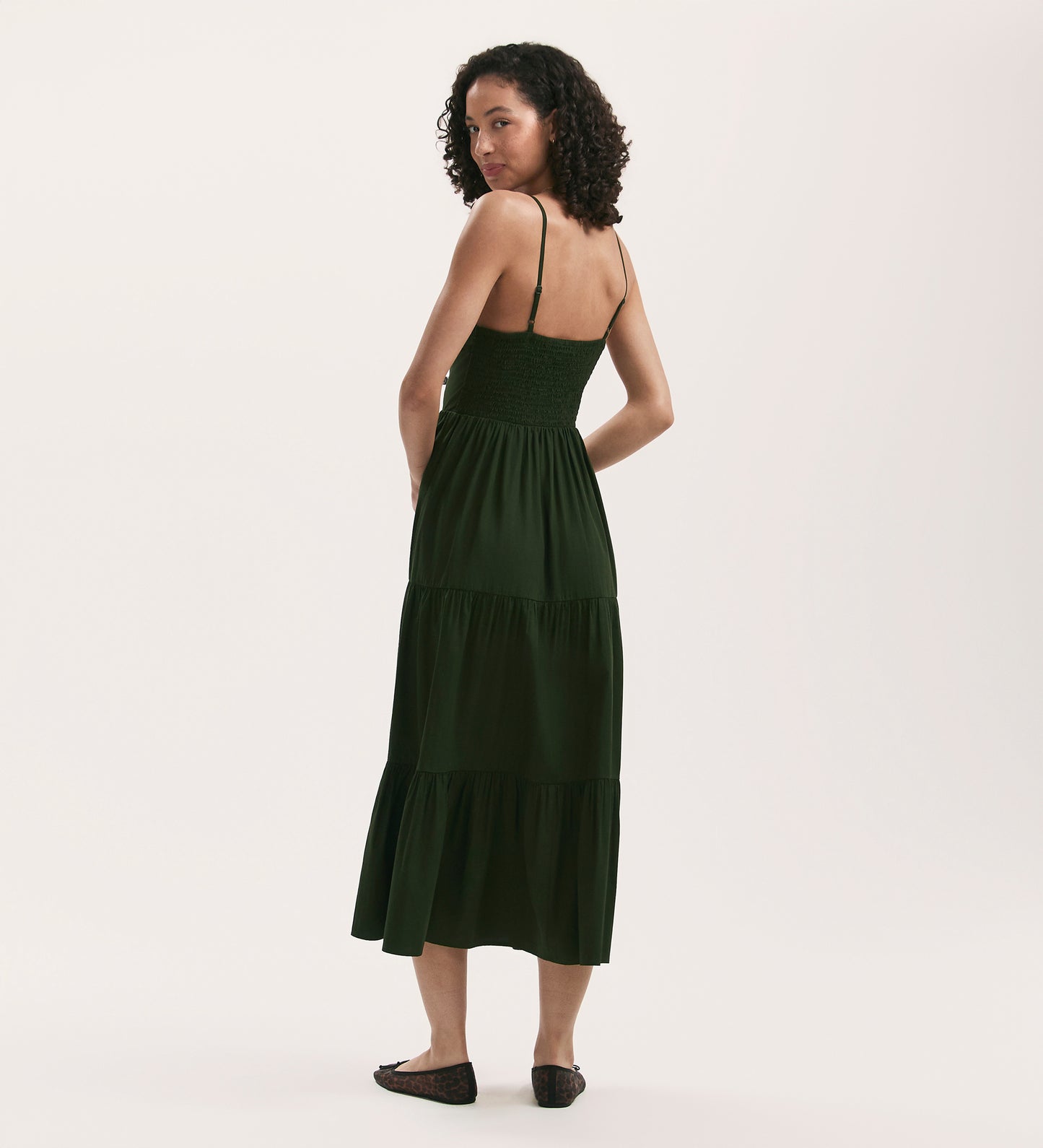 A woman with curly hair, facing backward and looking over her shoulder, wears the Emmie Dark Khaki Cotton Midi Dress with thin straps and a tiered skirt, paired with black flats against a plain white background.