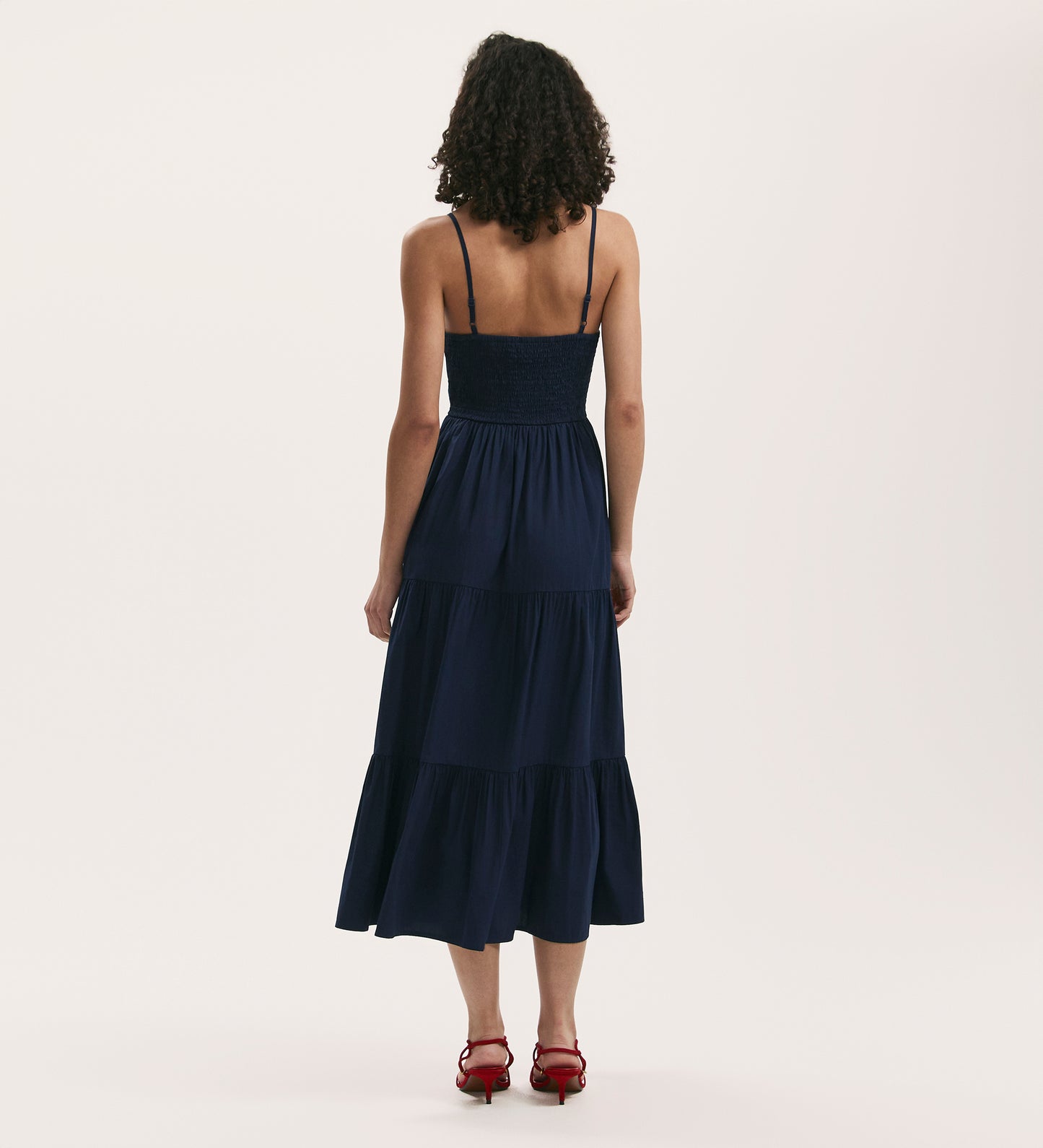 A woman with curly hair, in the Emmie Navy Cotton Midi Tiered Dress and red heeled sandals, stands facing away against a plain light background.