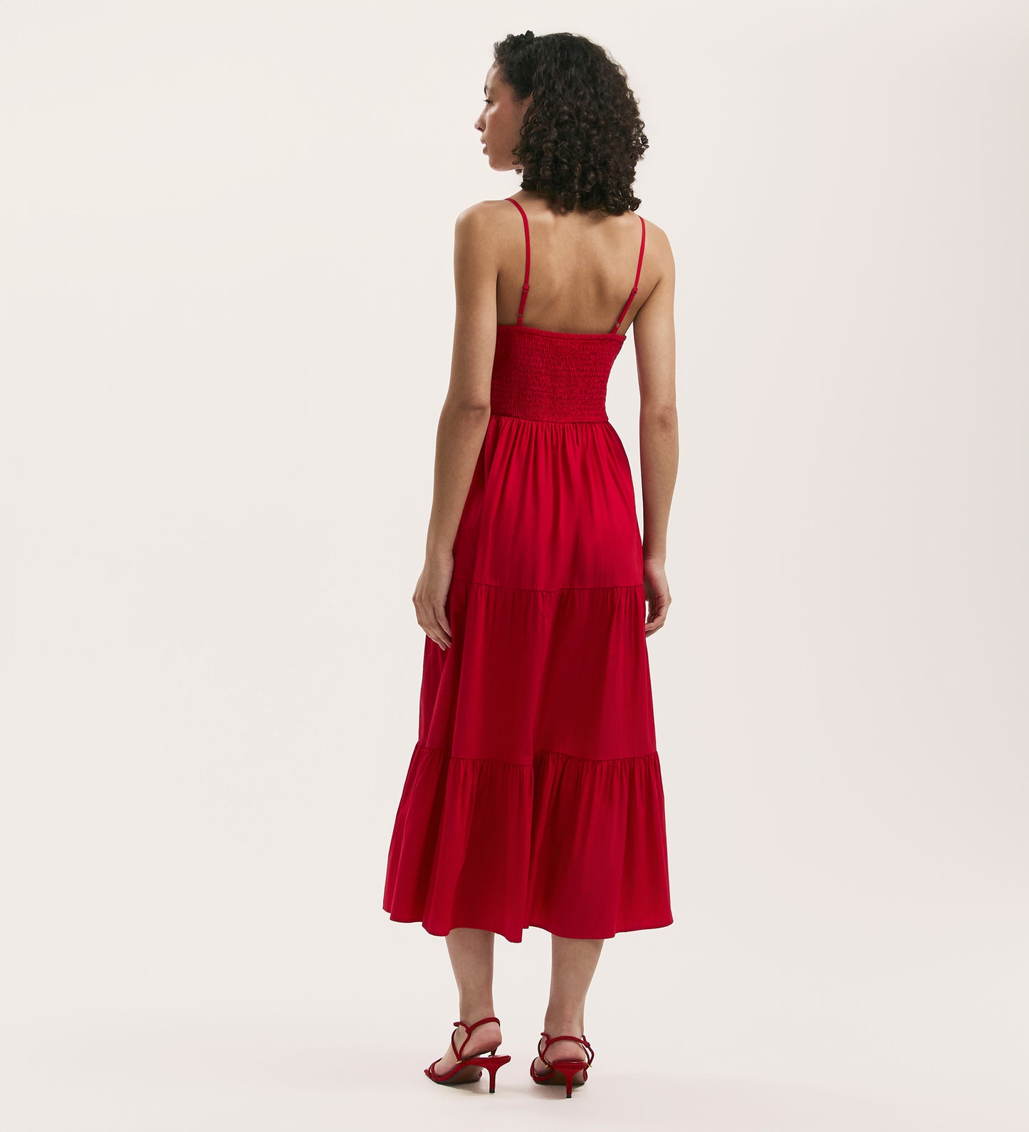 A woman with curly hair, seen from behind, wears the Emmie Red Cotton Midi Tiered Dress with thin straps and matching heeled sandals against a plain light background.