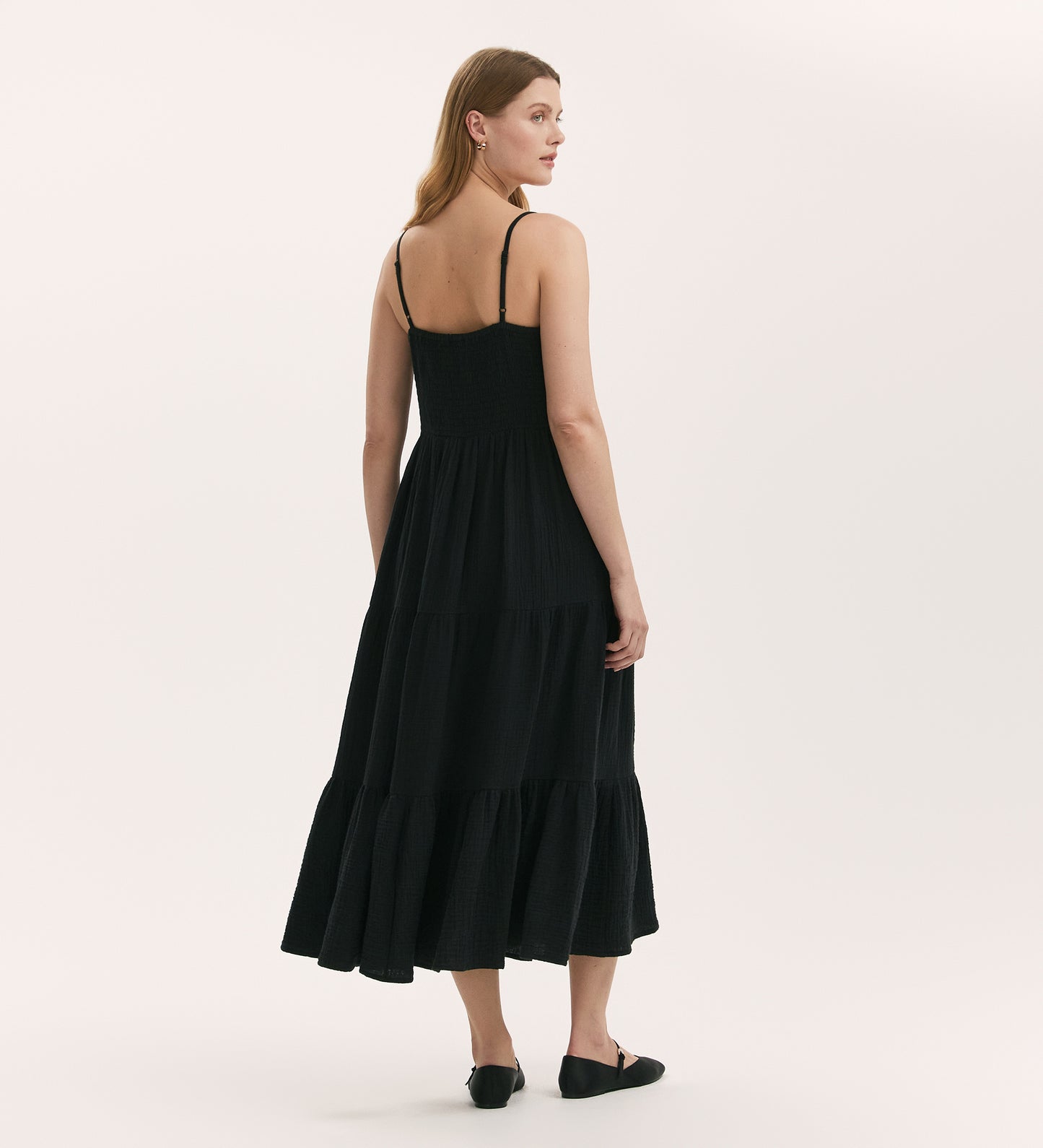 A woman with light brown hair stands facing away, looking over her shoulder, wearing the Emmie Black Cotton Midi Tiered Dress and black flats against a plain white background.