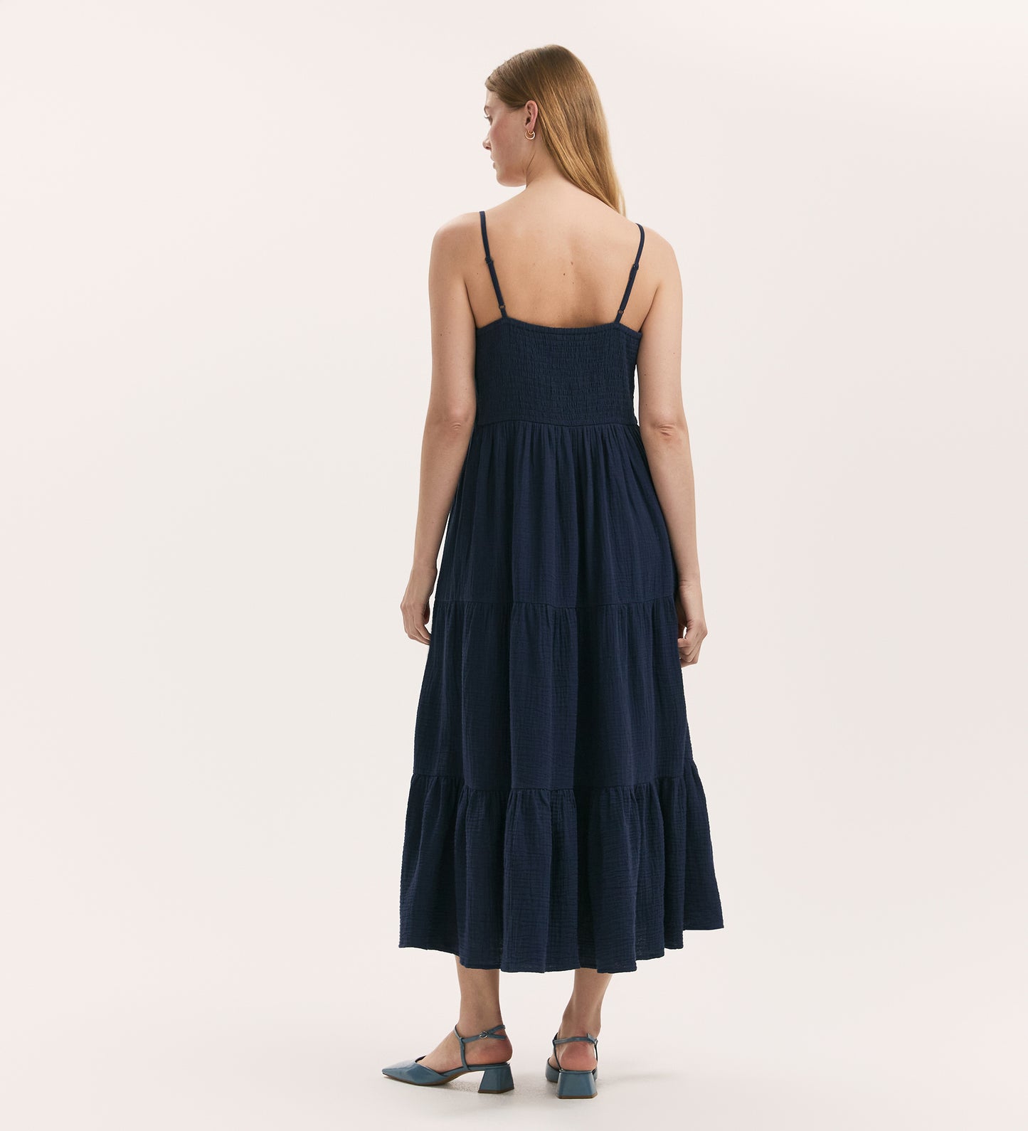 A woman with long, straight hair faces away, wearing the Emmie Navy Cotton Midi Tiered Dress with thin straps and a tiered skirt. She pairs it with light blue low-heeled shoes, set against a plain background.