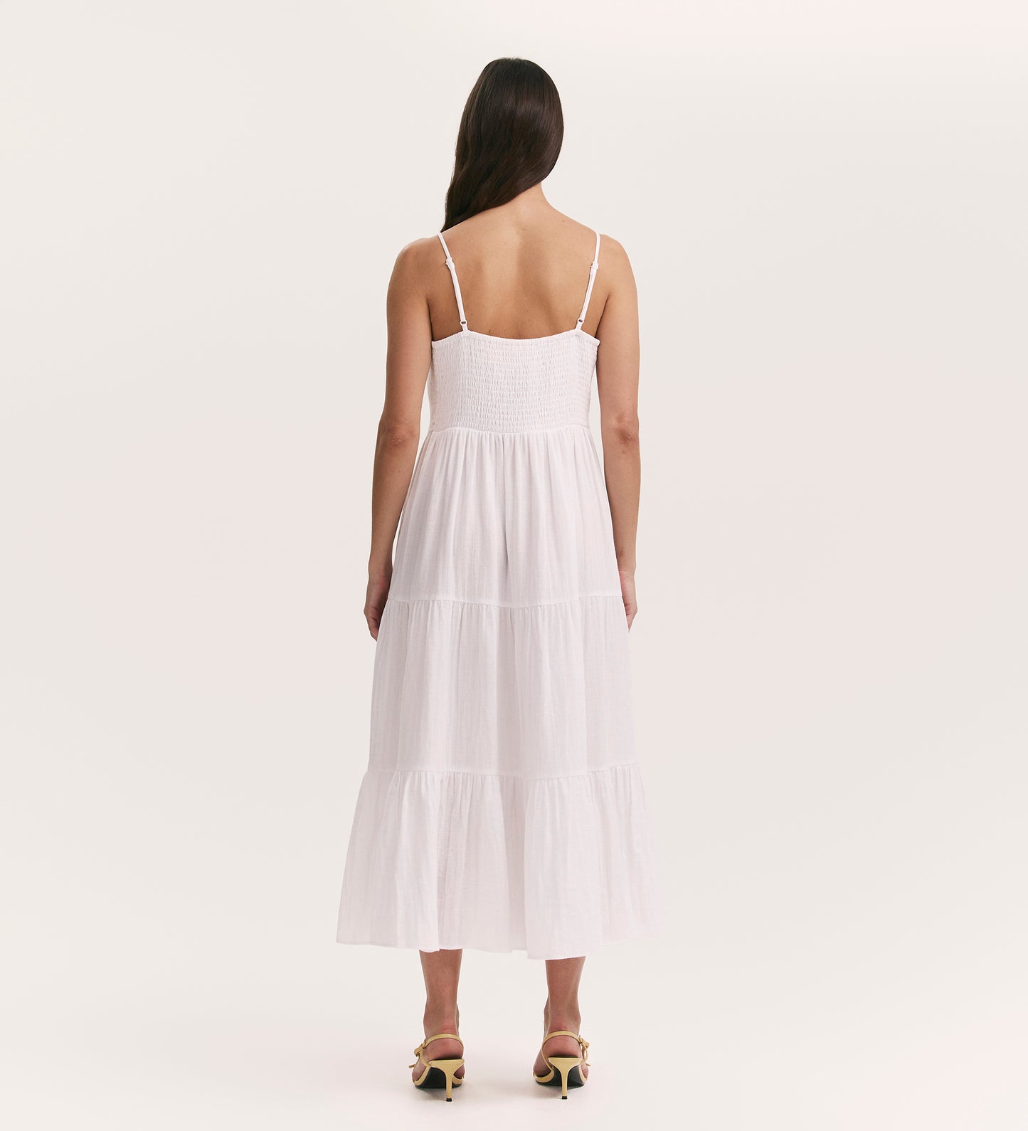 A woman with long brown hair, seen from behind, wears the Emmie White Cotton Midi Tiered Dress—sleeveless, with a tiered skirt in cheesecloth fabric—paired with heeled sandals, set against a plain light background.