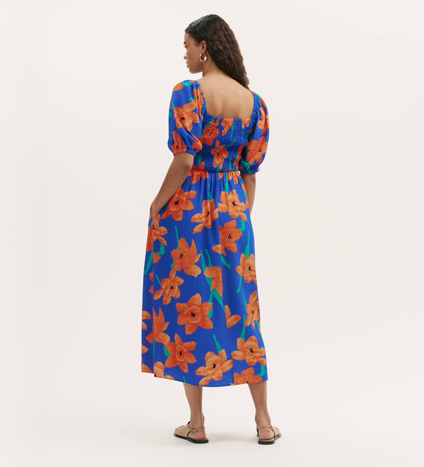 A woman faces away in the Enzo Blue Floral Midi Dress, featuring orange floral patterns, puffed short sleeves, a smocked bodice, and relaxed fit. She pairs it with flat sandals, hoop earrings, and long wavy hair—perfect for summer style.